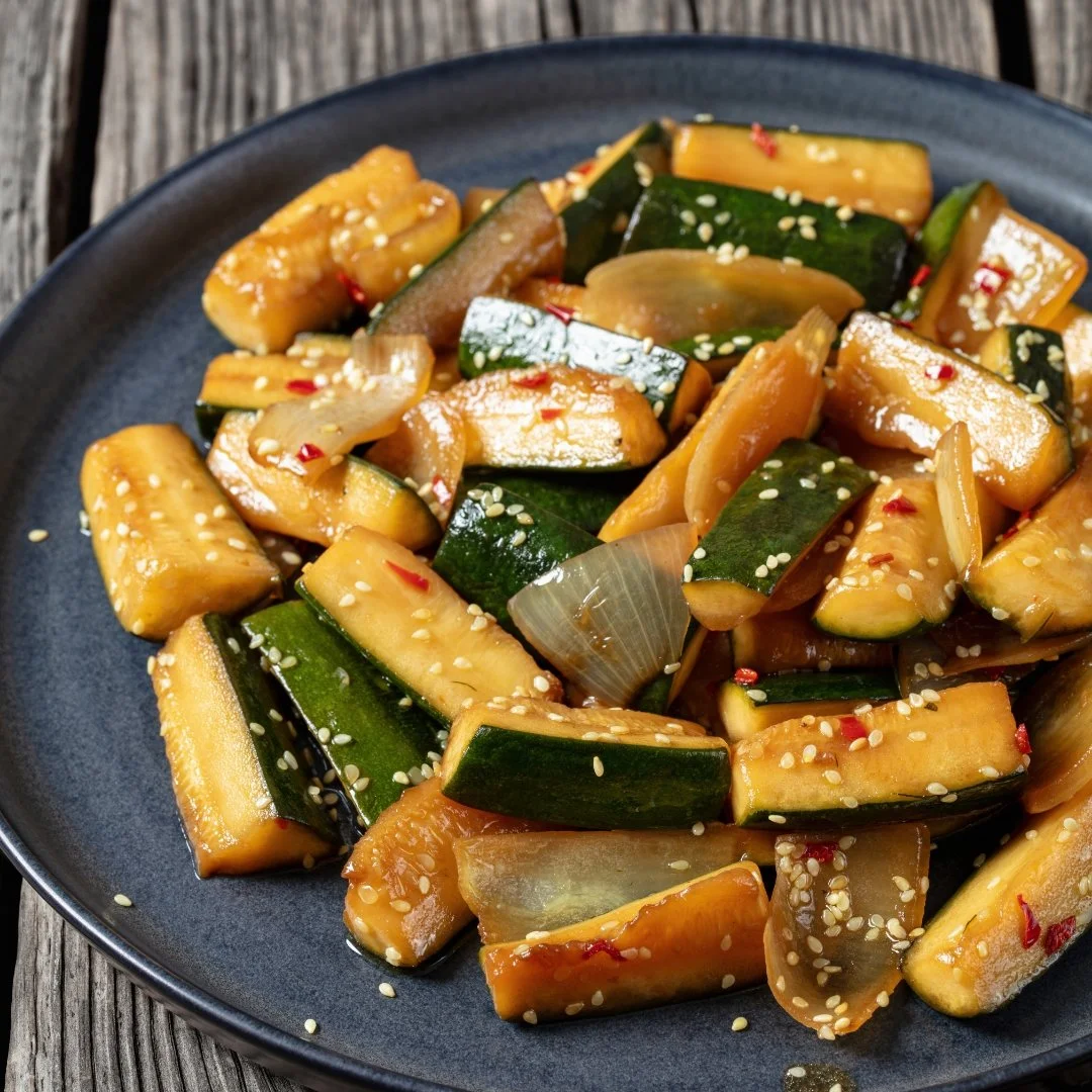 Cooked zucchini slices garnished with sesame seeds on a black plate, placed on a rustic wooden surface.