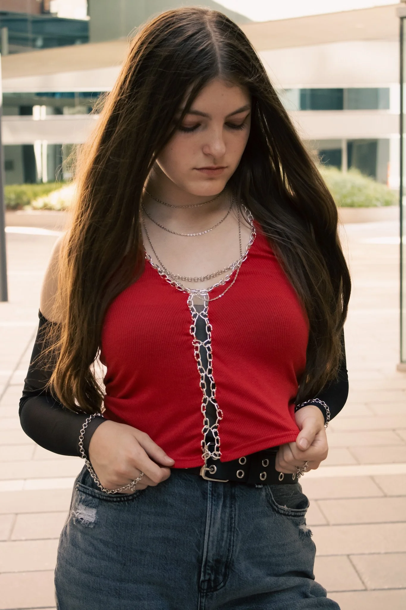 Young woman with long brown hair wearing a red top, black sleeve, and ripped blue jeans, looking down, outside urban background.
