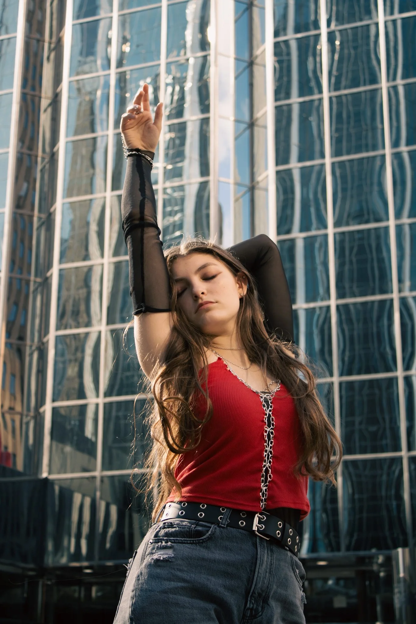 A young woman with long brown hair stands outdoors with her eyes closed, one arm raised above her head, against a backdrop of a modern glass building, wearing a red top, black mesh sleeves, and dark jeans.