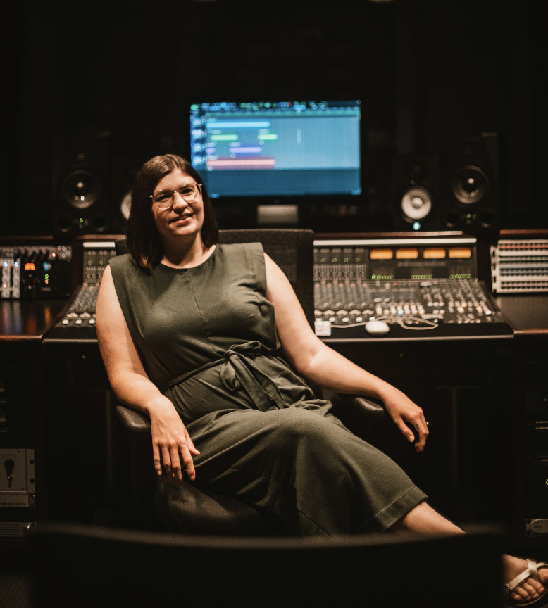 A woman in a seated position smiling in a recording studio surrounded by audio equipment with a computer screen displaying a digital audio workstation in the background.