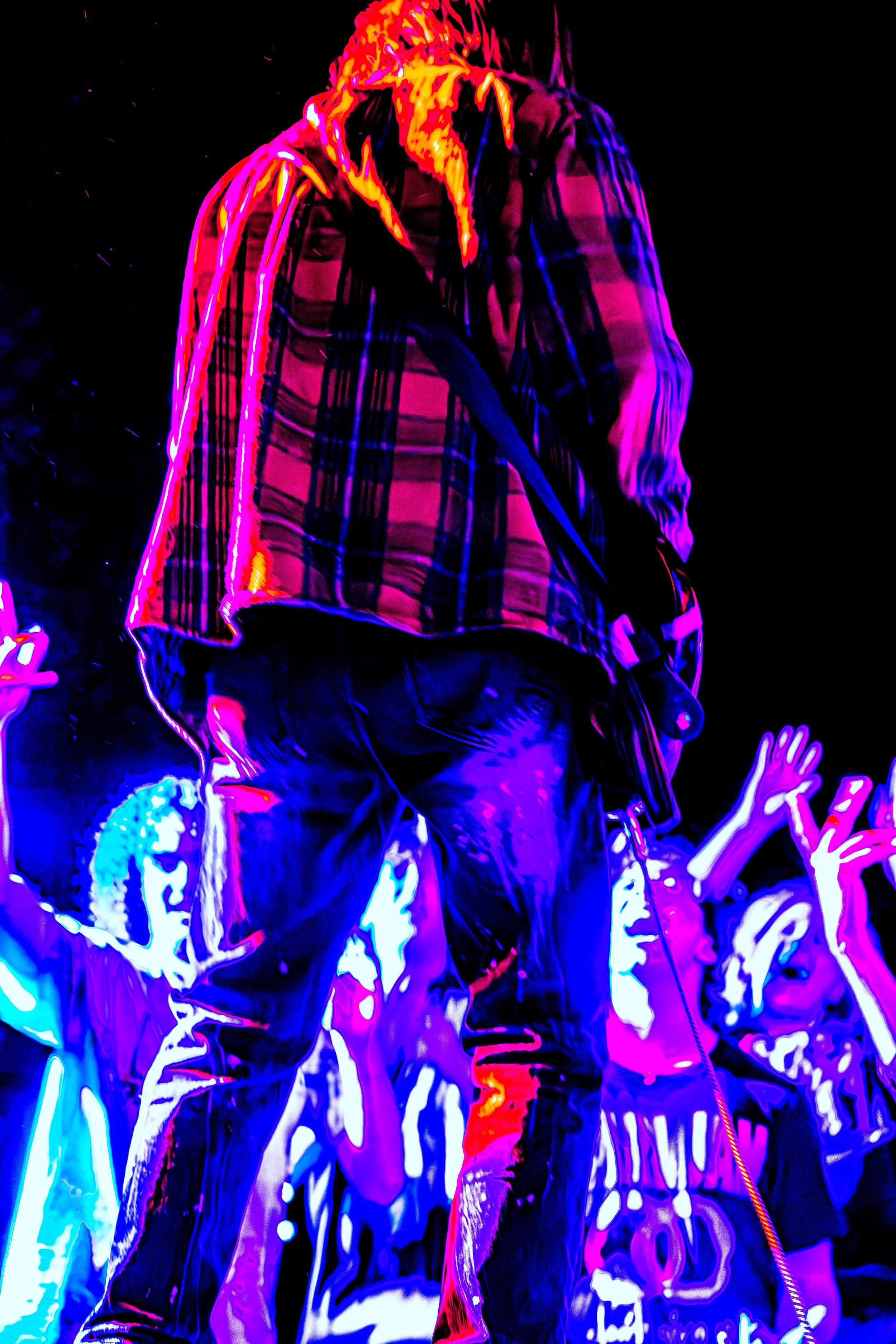 A performer on stage with colorful neon lighting, wearing a plaid shirt and jeans, facing away from the camera, with a crowd of raised hands behind.