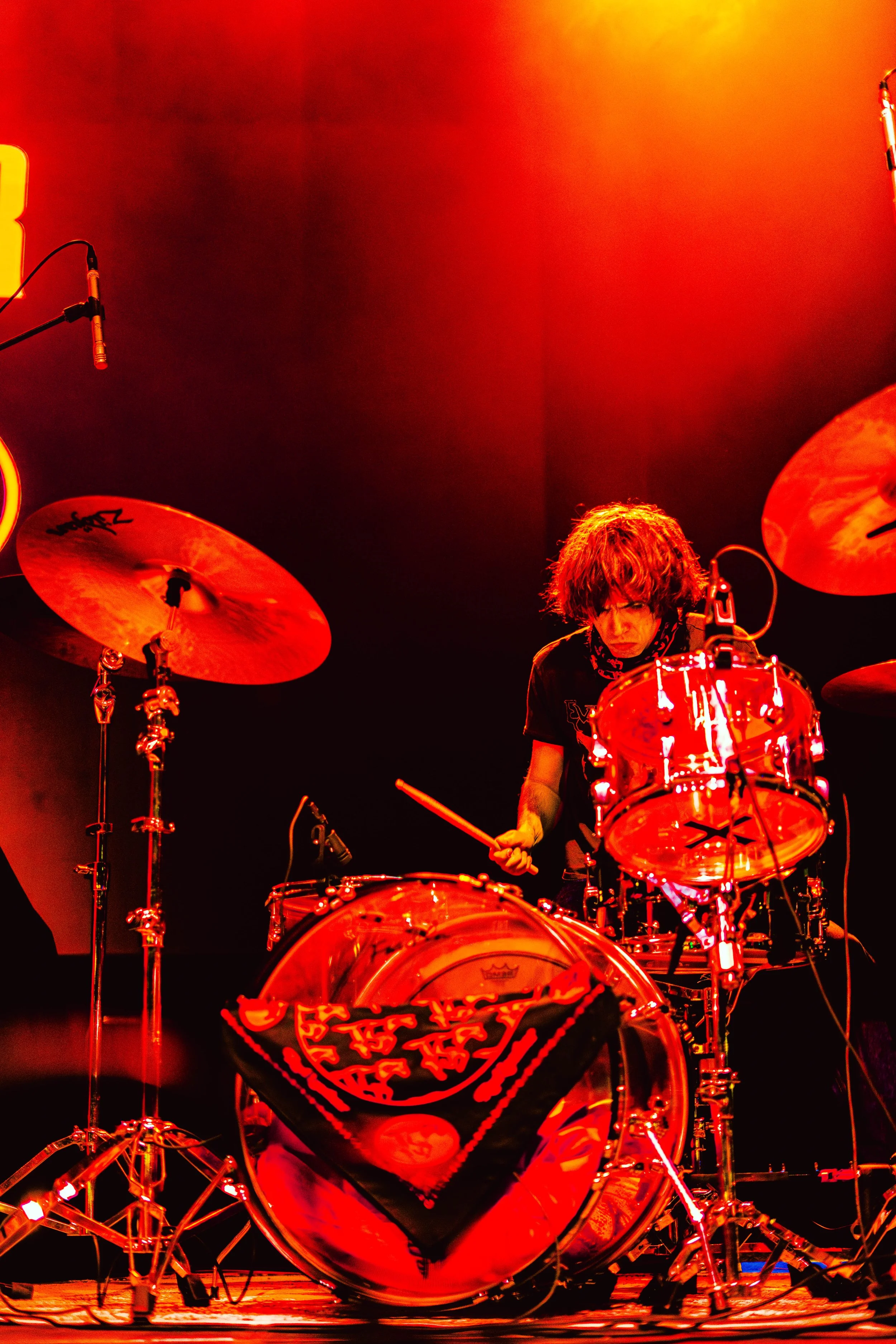 A drummer performing on stage, illuminated by red and orange stage lights, surrounded by his drum kit and cymbals.