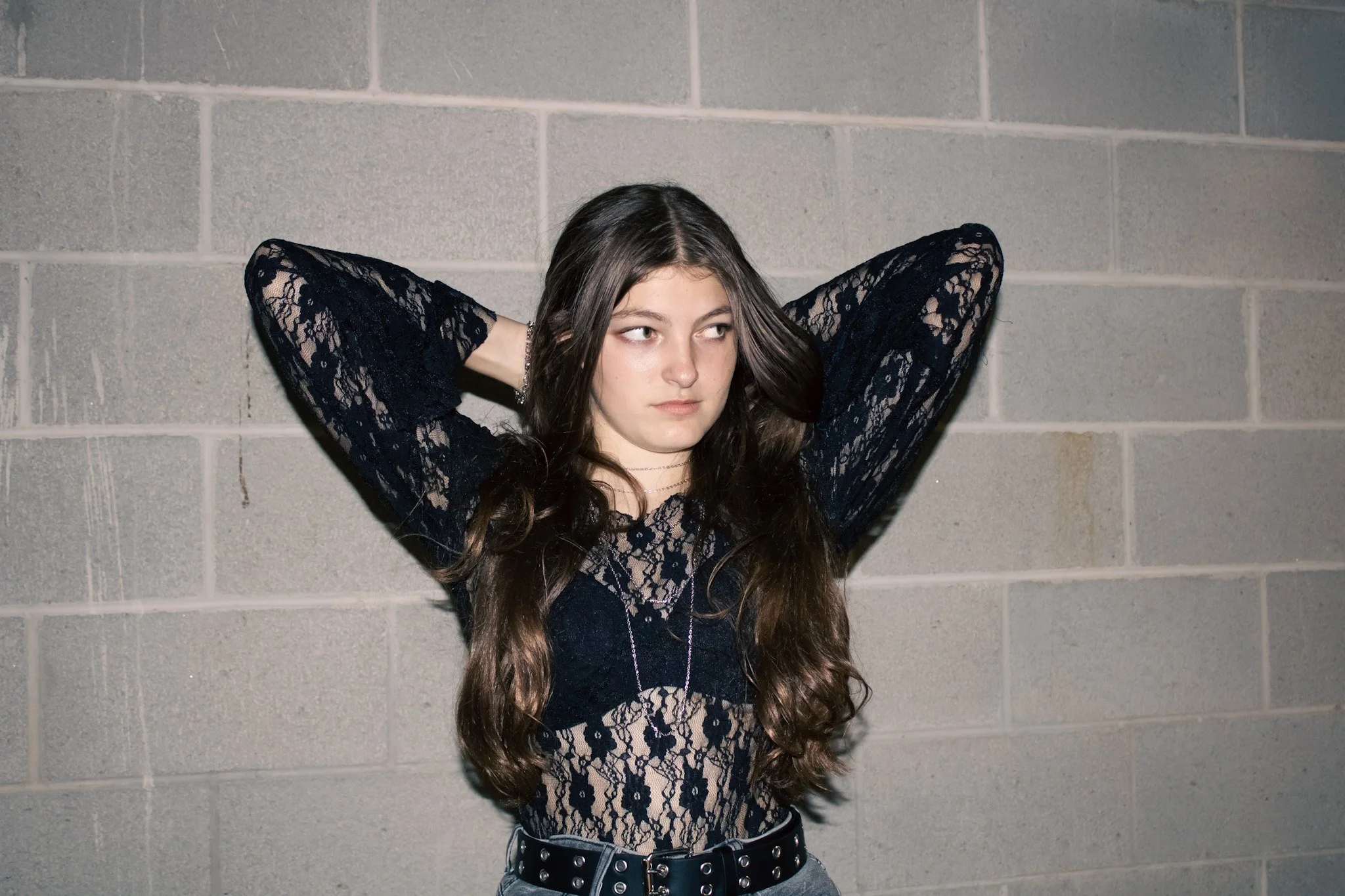 Young woman with long, wavy brown hair standing against a gray brick wall, wearing a black lace top, with arms raised and hands behind her head, looking to the side.