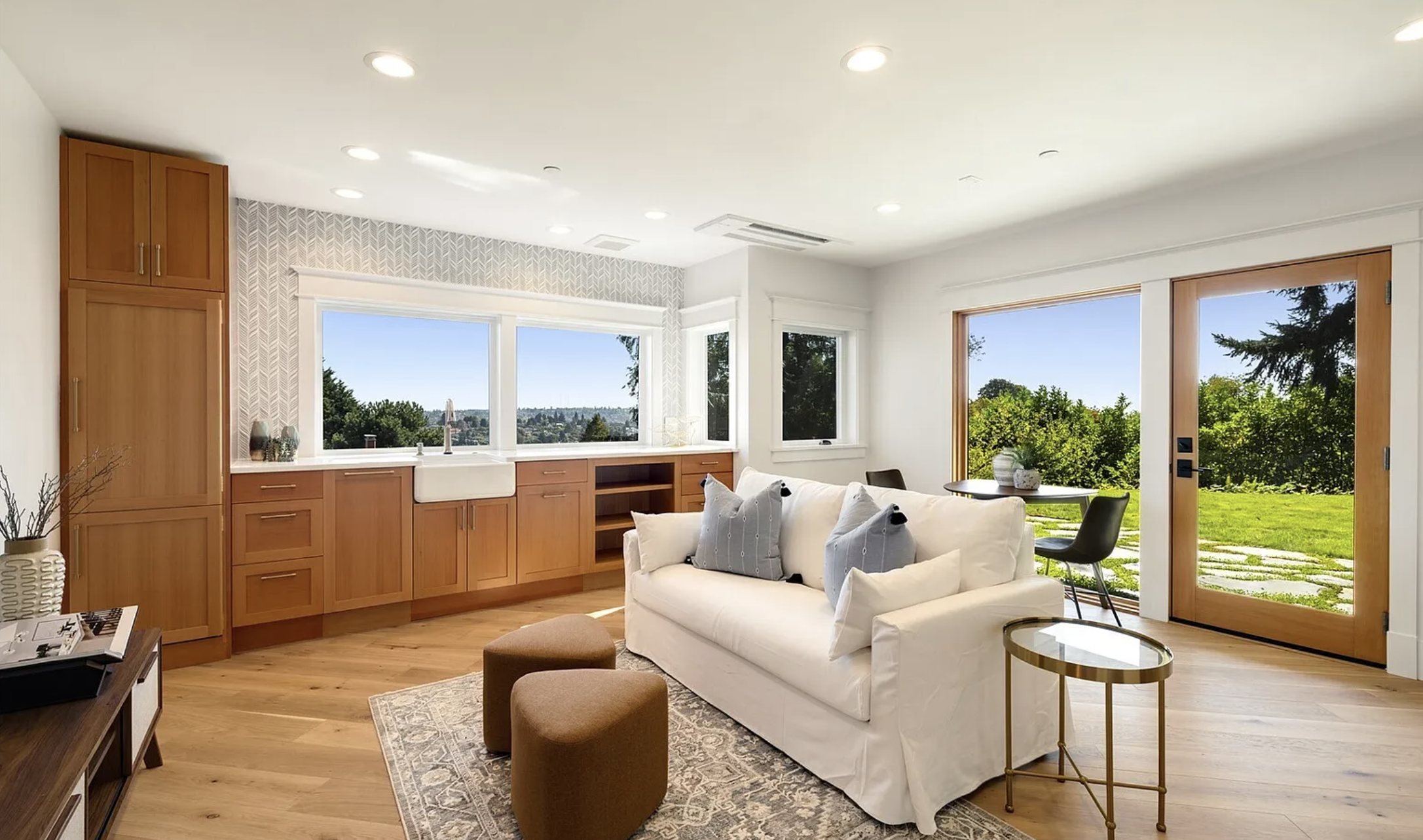 Living room with white sofa, blue pillows, wooden floor, large windows, sliding glass door opening to outdoor green yard, kitchen with wooden cabinets, and view of trees and blue sky