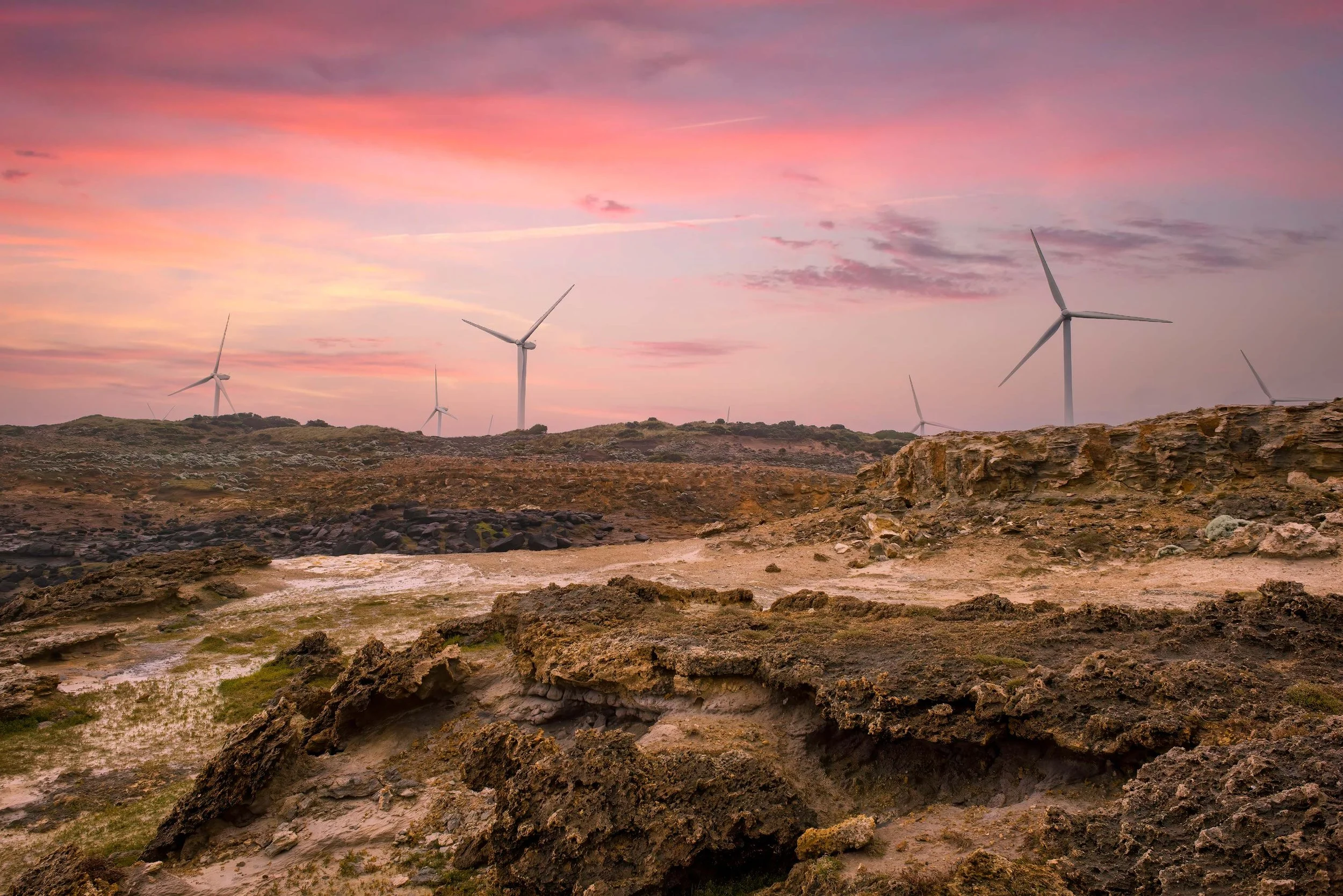 Wind turbines on a rocky terrain under a pink and purple sunset sky.