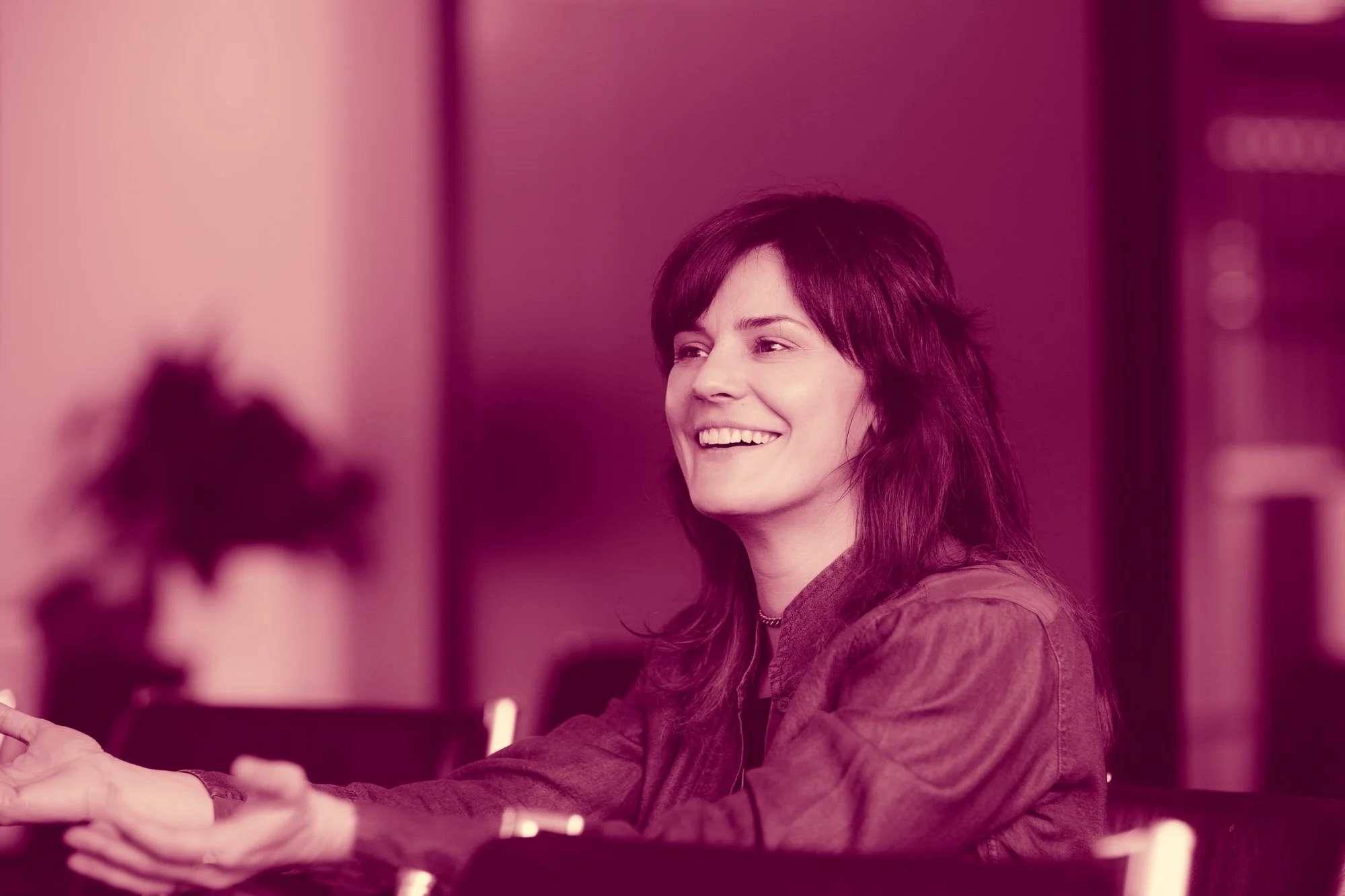 A woman with dark hair smiling and looking to her right, sitting at a table with her hands extended forward.