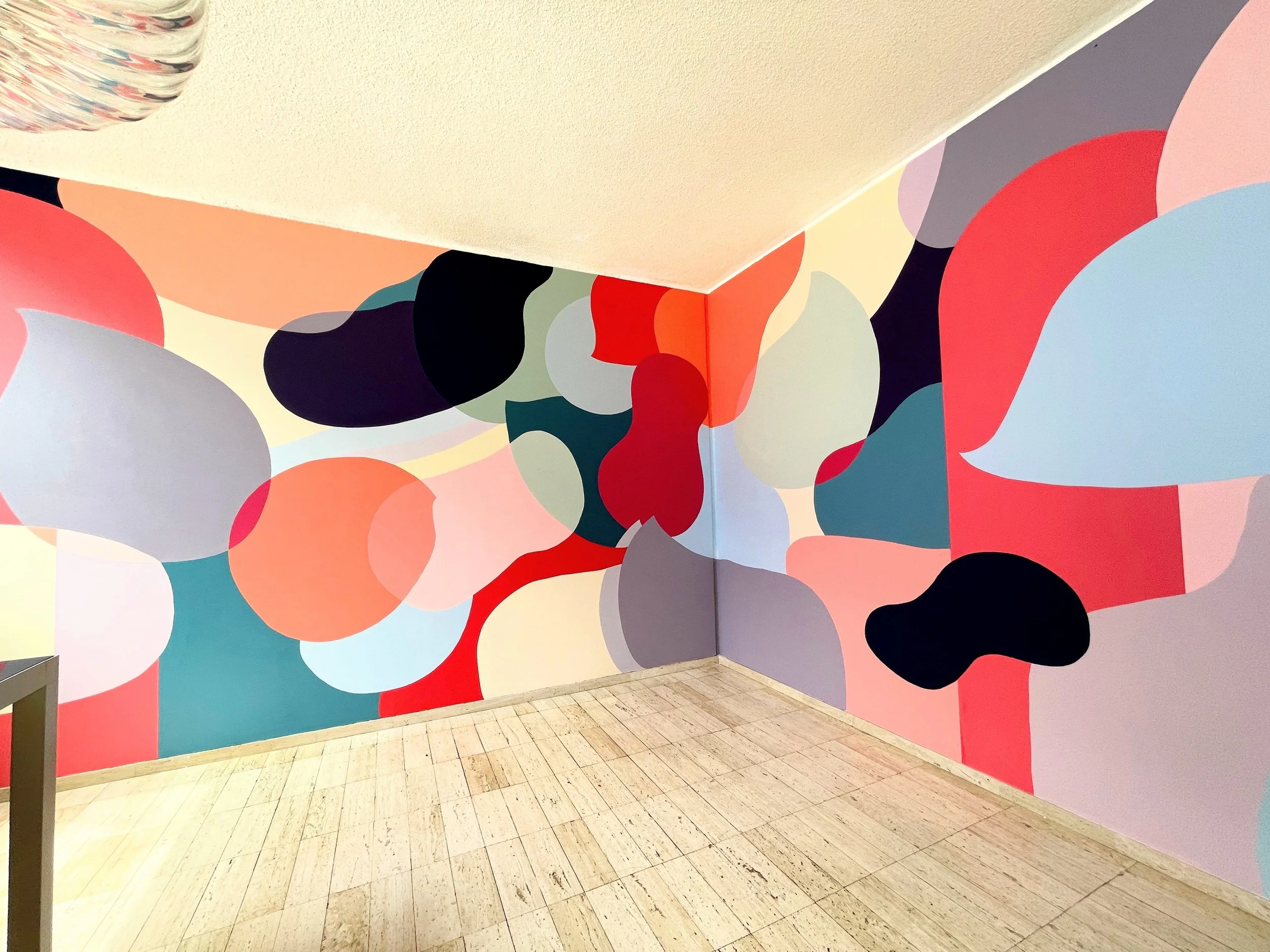 Colorful abstract mural with organic shapes in pink, red, black, gray, green, blue, purple, and peach on a corner wall of a room with a beige tiled floor.