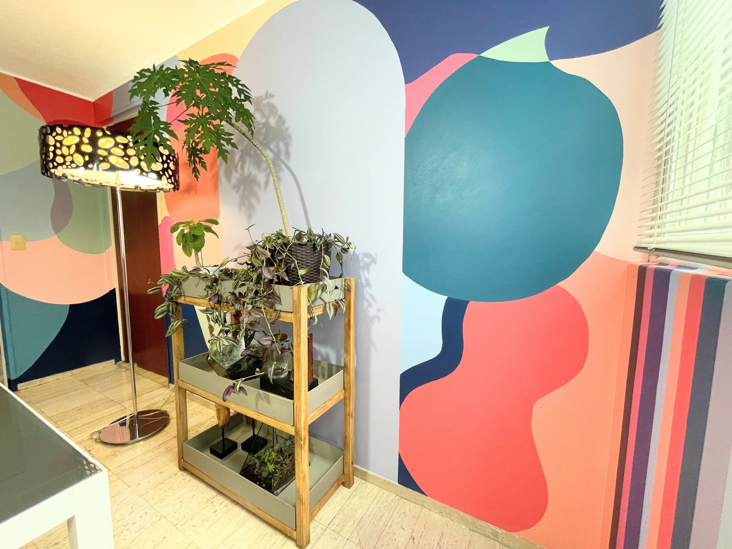 Interior space with a colorful abstract wall mural featuring large shapes in pink, green, blue, and white. A wooden plant stand with several potted plants, one climbing with purple-tinged leaves, is positioned in front of the mural. To the left, a ta