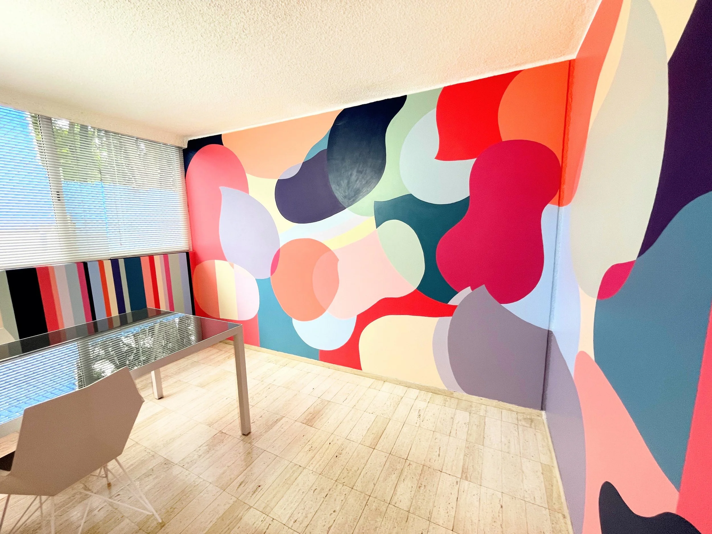 Interior of a room with a colorful abstract mural on the wall, a metal table, and white chairs; window with blinds letting in natural light.