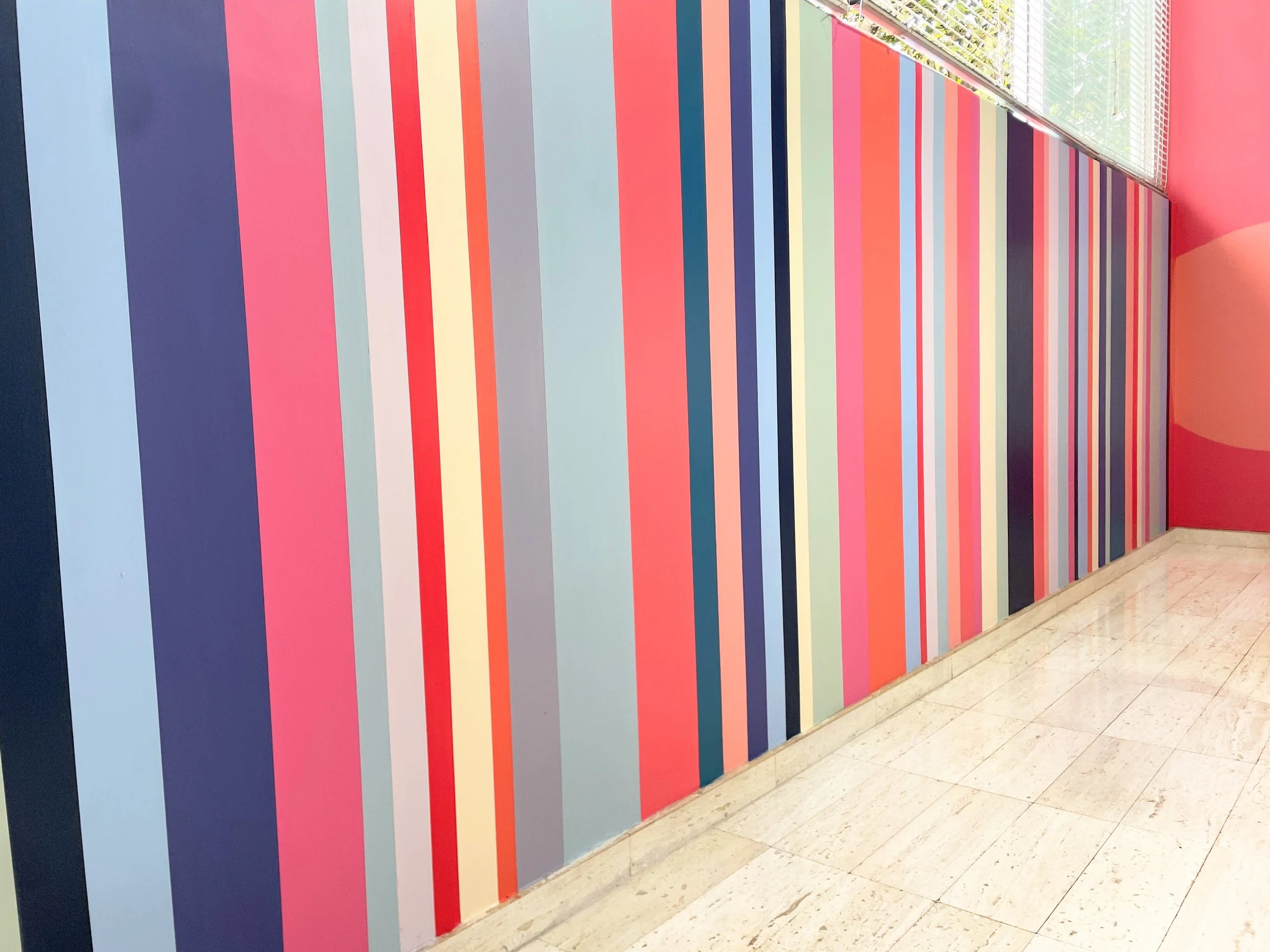 Colorful vertical stripes on a wall in shades of blue, pink, yellow, orange, gray, and purple, with a window at the top right allowing natural light to illuminate the area.