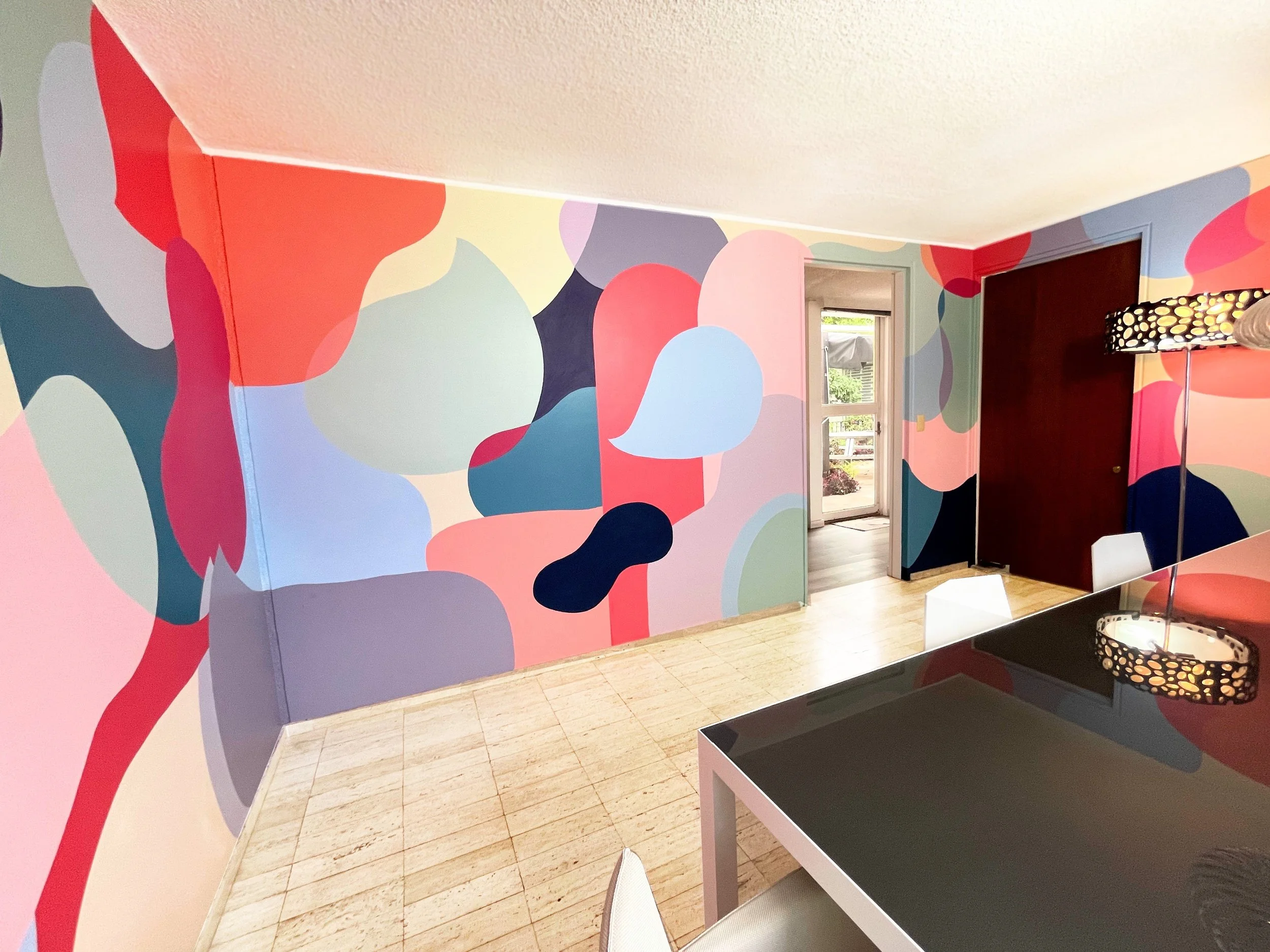Room with colorful abstract mural on walls featuring shapes in red, pink, purple, blue, green, and black. Furniture includes a black table, white chair, and floor lamp with a black dotted shade. There is a doorway leading outside and a window.