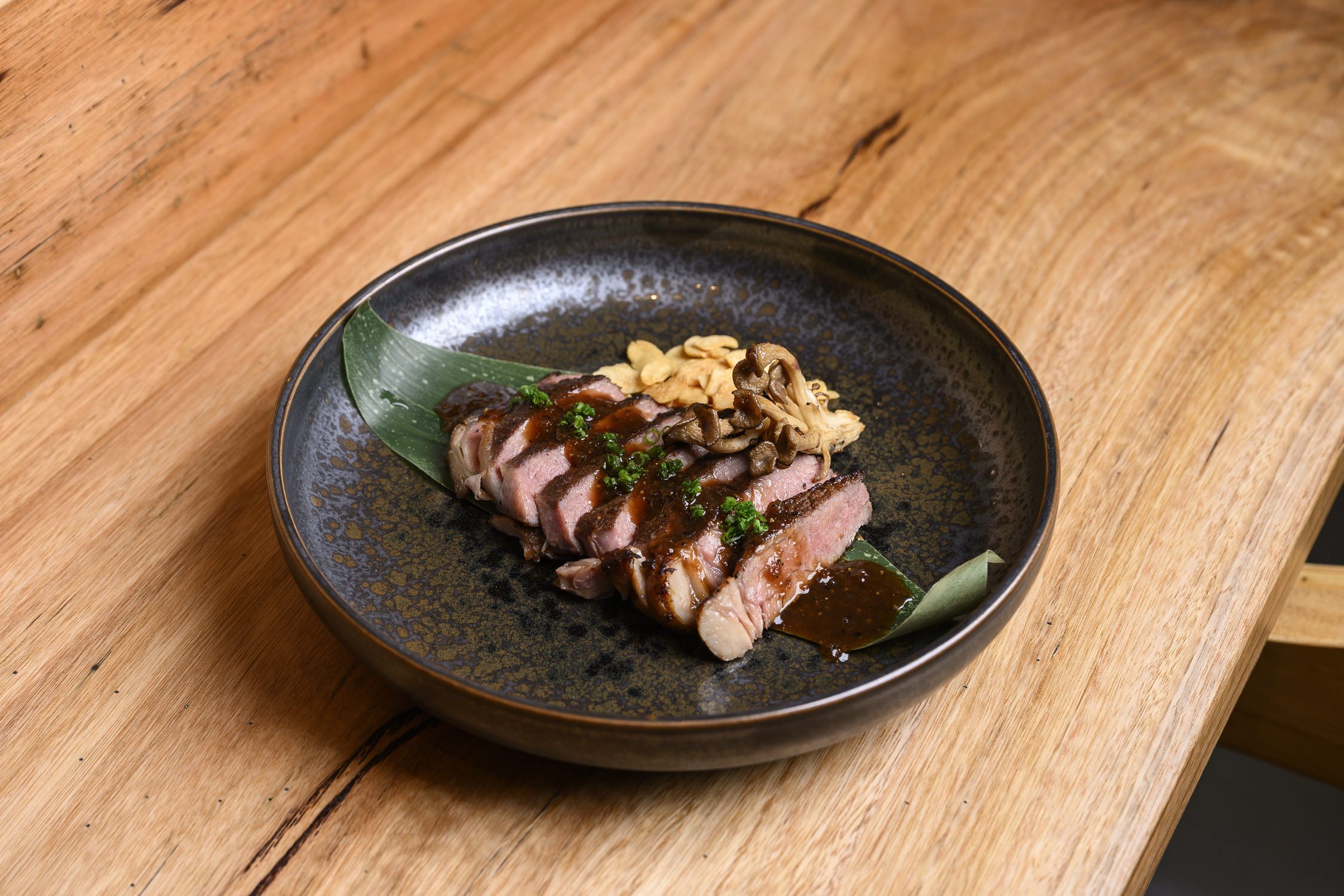 Slices of seared tuna topped with sauce, chopped green onions, and mushrooms on a black plate with a green leaf garnish.
