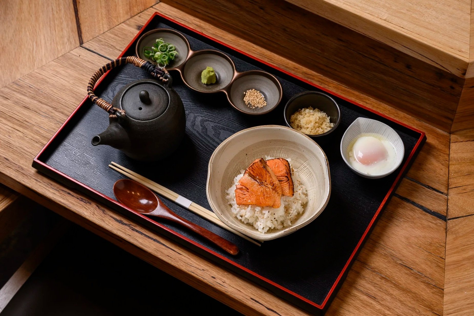 Japanese meal served on a black tray with a bowl of rice topped with grilled fish, a soft-boiled egg, a teapot, and small dishes of green onions, wasabi, sesame seeds, grated radish, and bonito flakes.