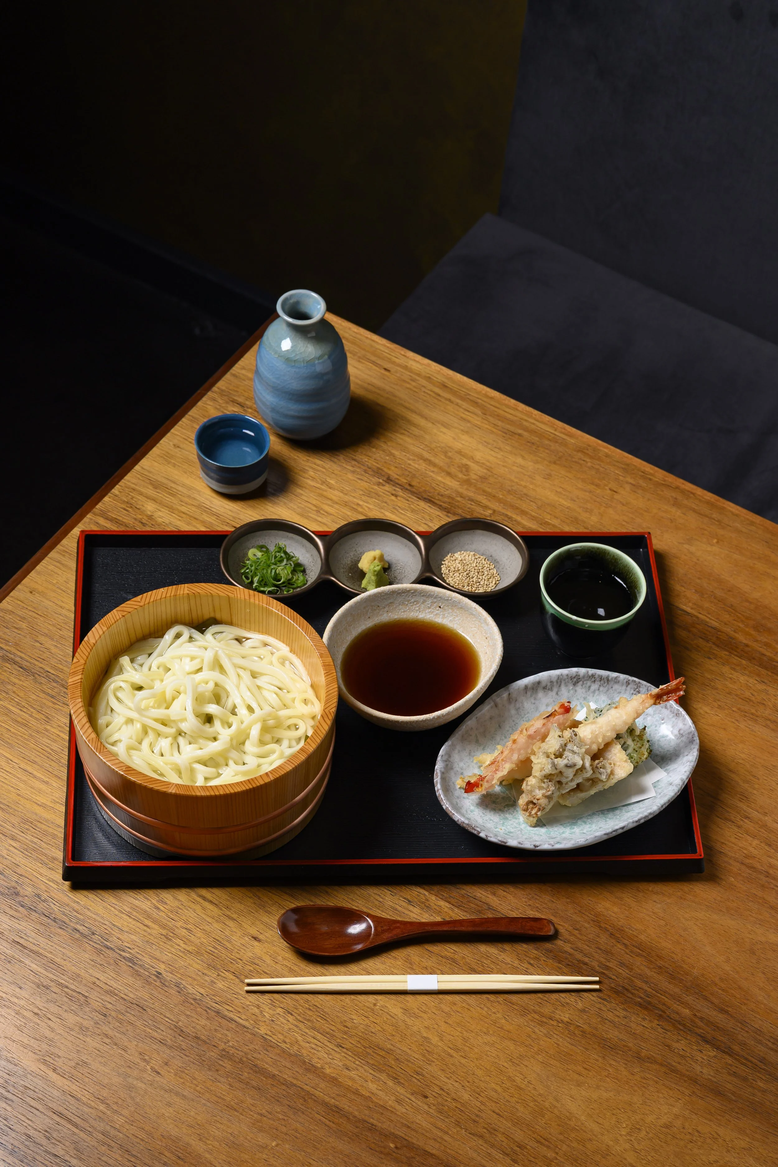 A traditional Japanese meal with a bowl of udon noodles, tempura, dipping sauce, soy sauce, and condiments, arranged on a black tray on a wooden table.