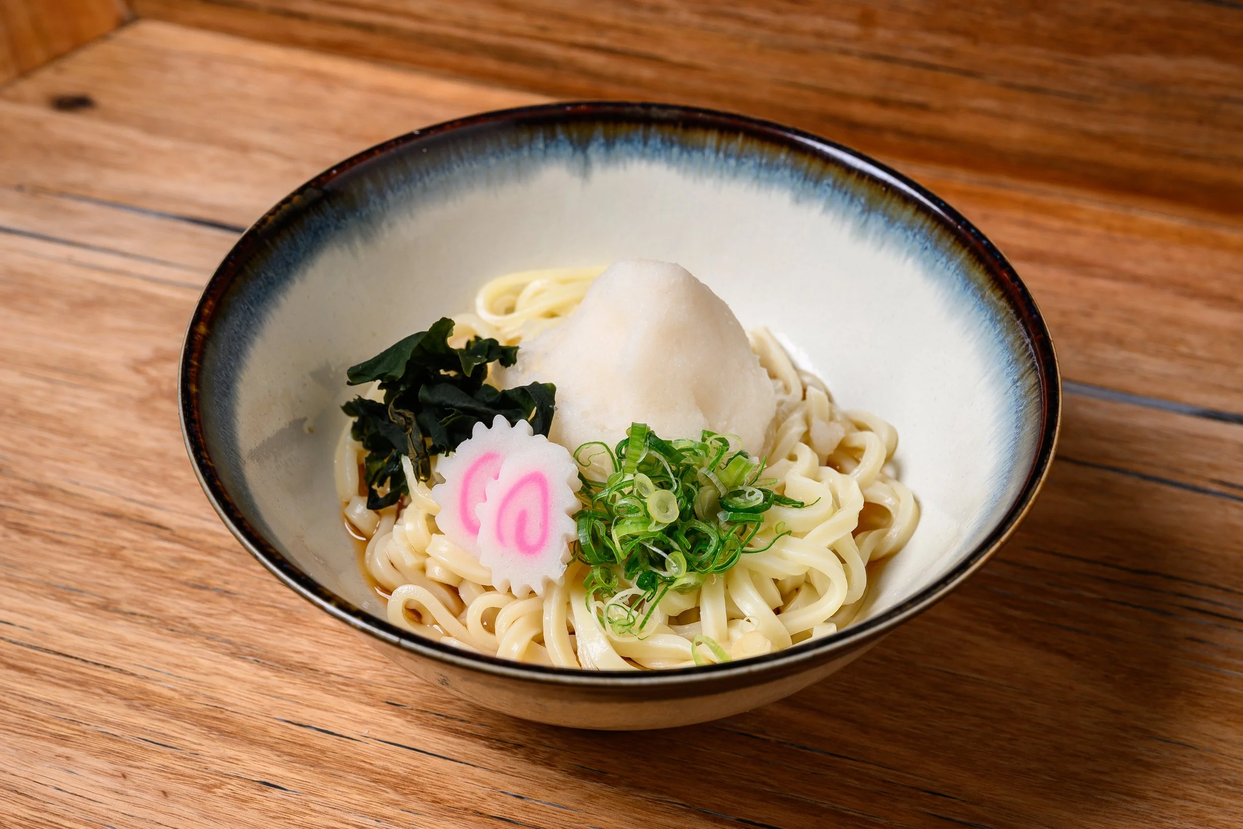 Bowl of Japanese ramen with noodles, topped with grated daikon radish, seaweed, chopped green onions, and a fish cake with pink swirls, served on a wooden table.