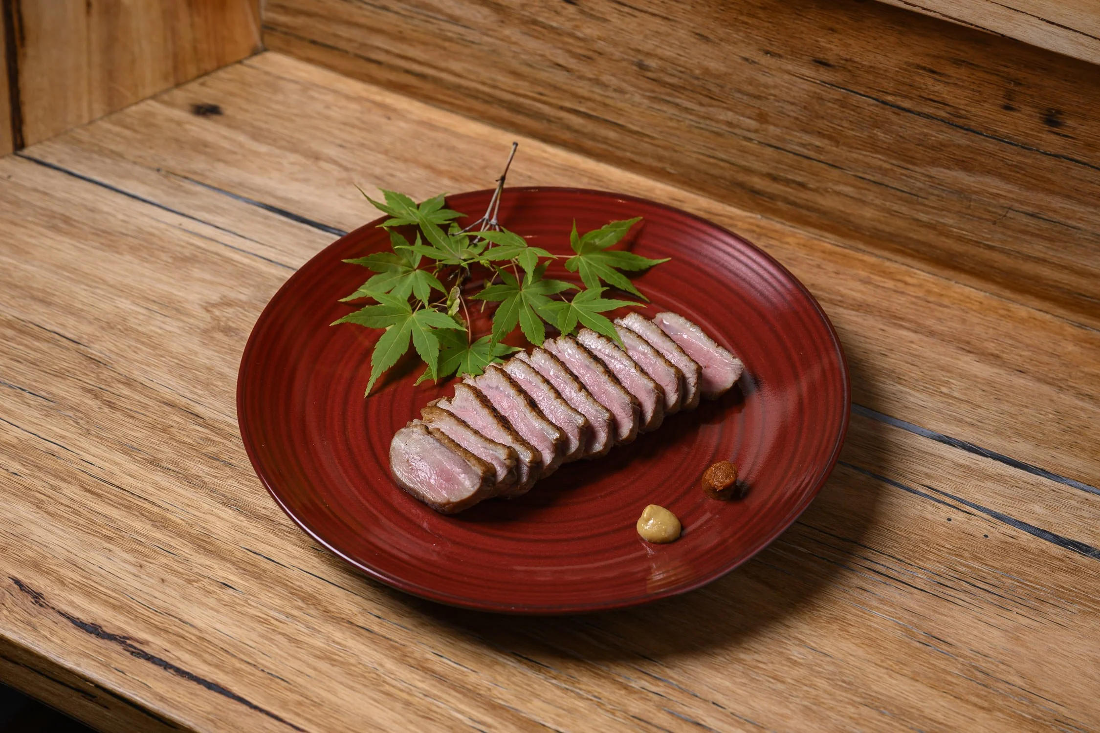 Slices of seared tuna with green maple leaves and mustard, wasabi, and sauce on a red plate on a wooden surface.