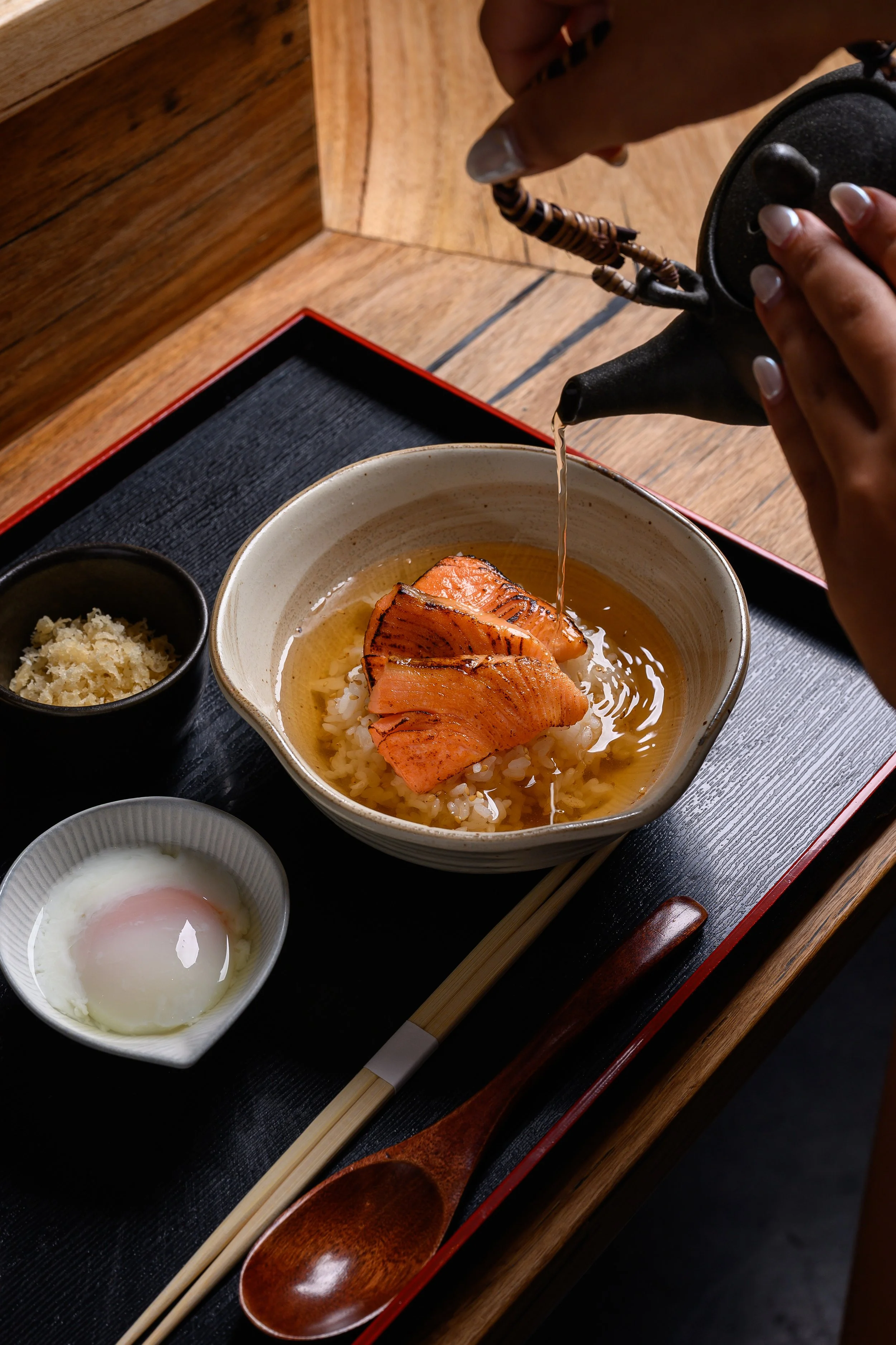 A bowl of rice topped with grilled salmon, being poured with broth. There is a soft-boiled egg, grated ginger, and a small bowl of sauce or seasoning on a black tray with wooden chopsticks and spoon.