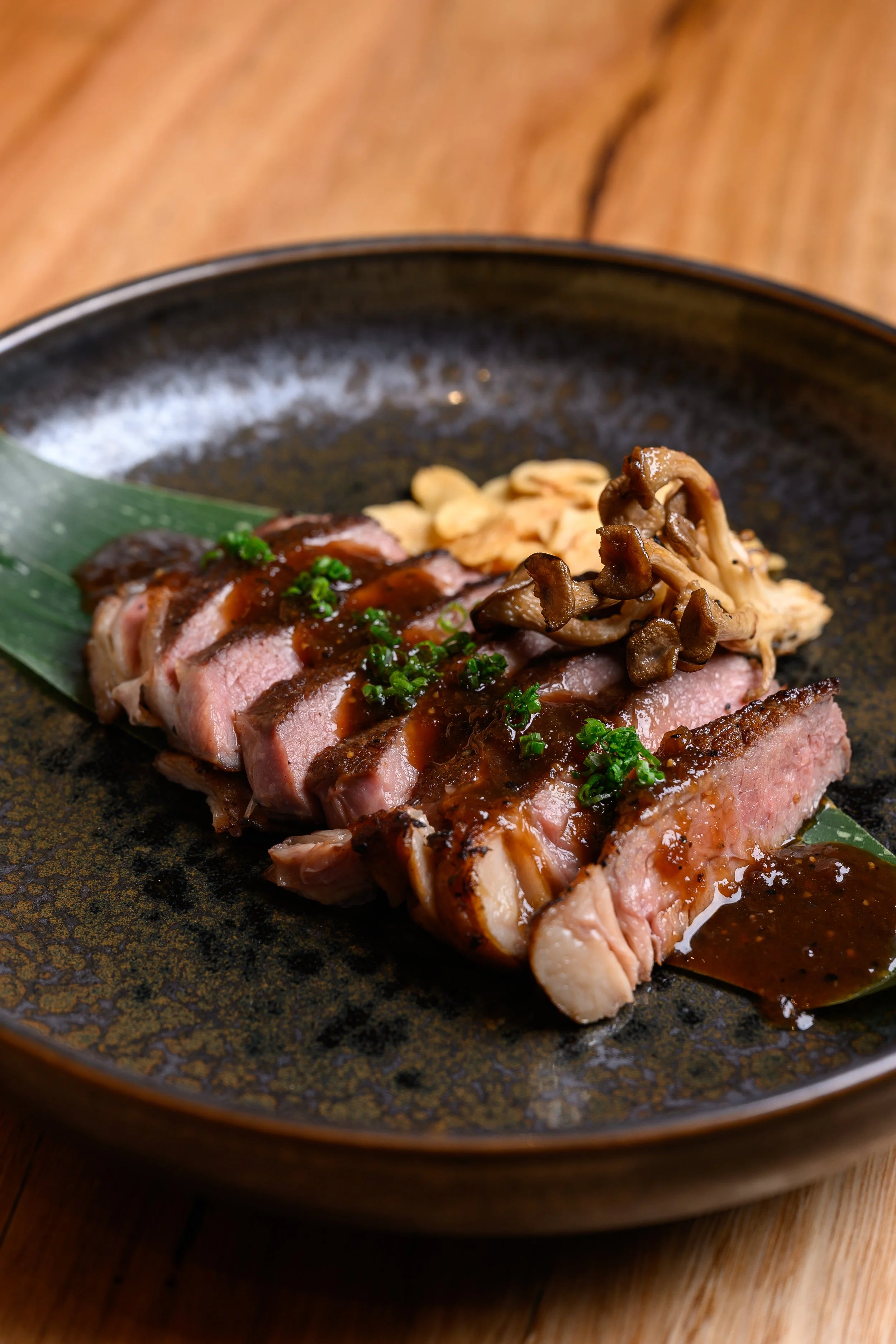 Sliced cooked steak with sauce, topped with chopped chives, served on a black plate with mushrooms in the background.