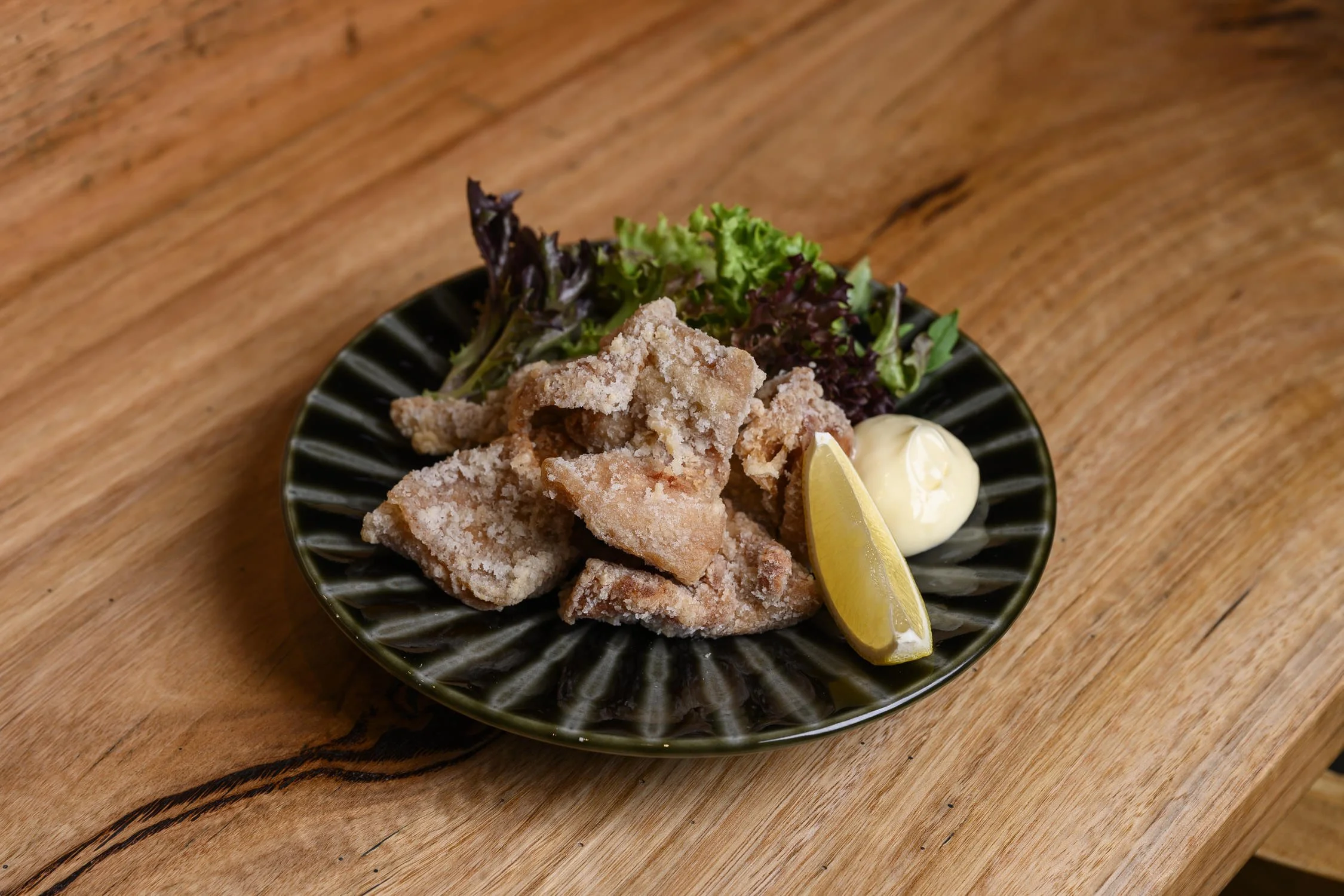 Fried fish served with lemon wedge, a dollop of mayonnaise, and mixed greens on a black plate.