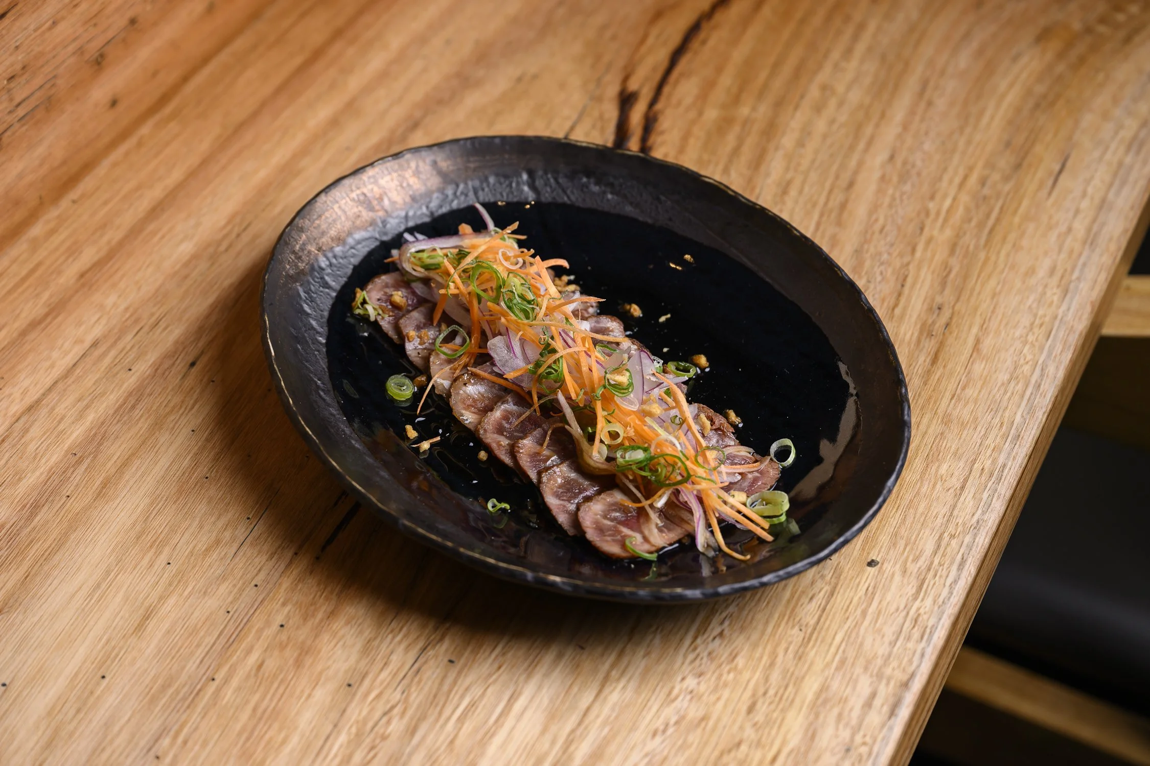 Sliced raw fish garnished with sliced green onions, shredded carrots, and thinly sliced radishes on a black plate.
