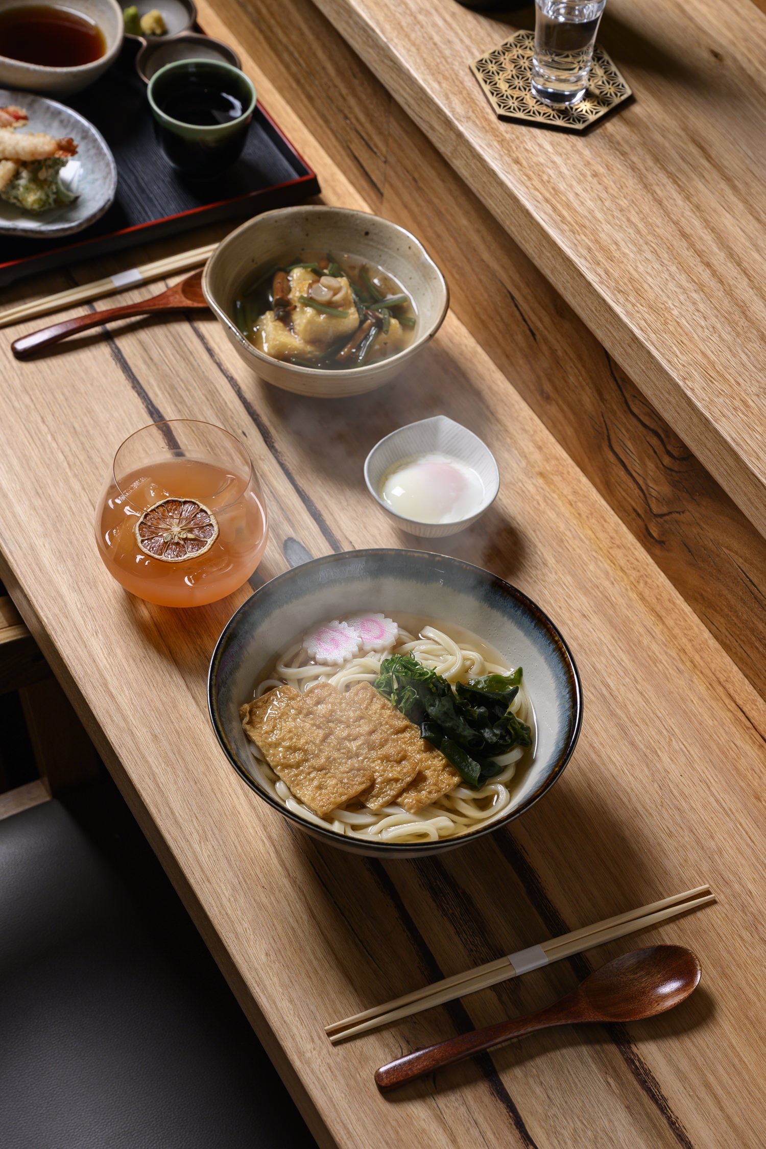 A Japanese meal on a wooden table featuring a bowl of udon noodles with tofu, greens, and fish cake, a small bowl of soft-boiled egg, a glass of iced tea with lemon, a bowl of hot soup with vegetables and tofu, and a tray with dipping sauces and tempura, accompanied by chopsticks and a wooden spoon.