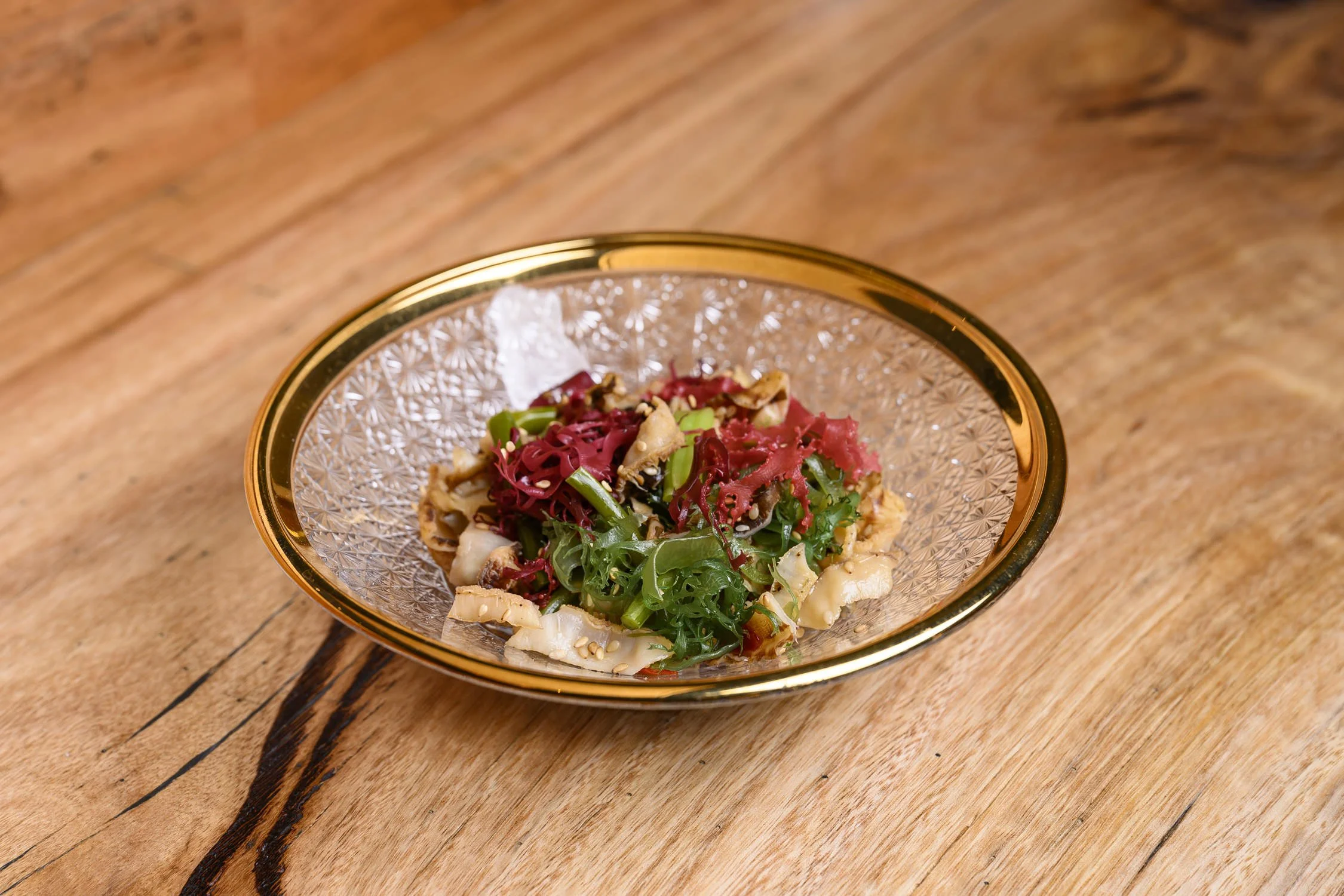 A decorative glass bowl with a gold rim filled with a colorful salad of mixed greens, red cabbage, and chopped vegetables on a wooden table.