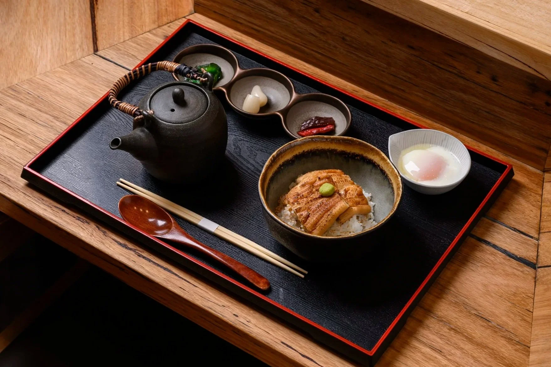 Japanese meal with a bowl of rice topped with cooked eel, a small dish with a soft-boiled egg, a teapot, and small dishes with pickles and sauces on a black tray.