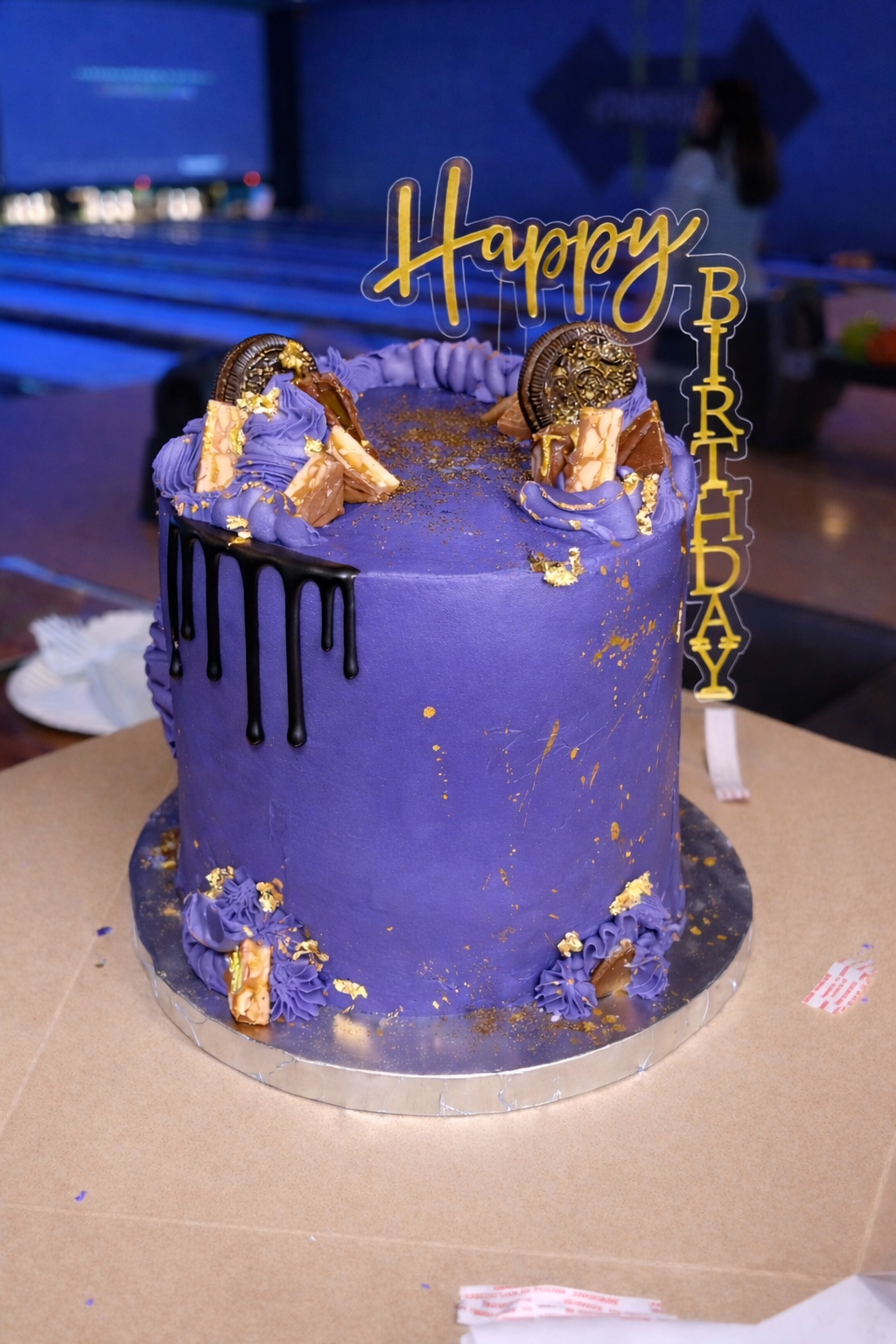 A tall purple birthday cake with black drip decorations, gold accents, Oreo cookies, and purple and gold frosting decorations, topped with a 'Happy Birthday' topper, on a round silver cake board.