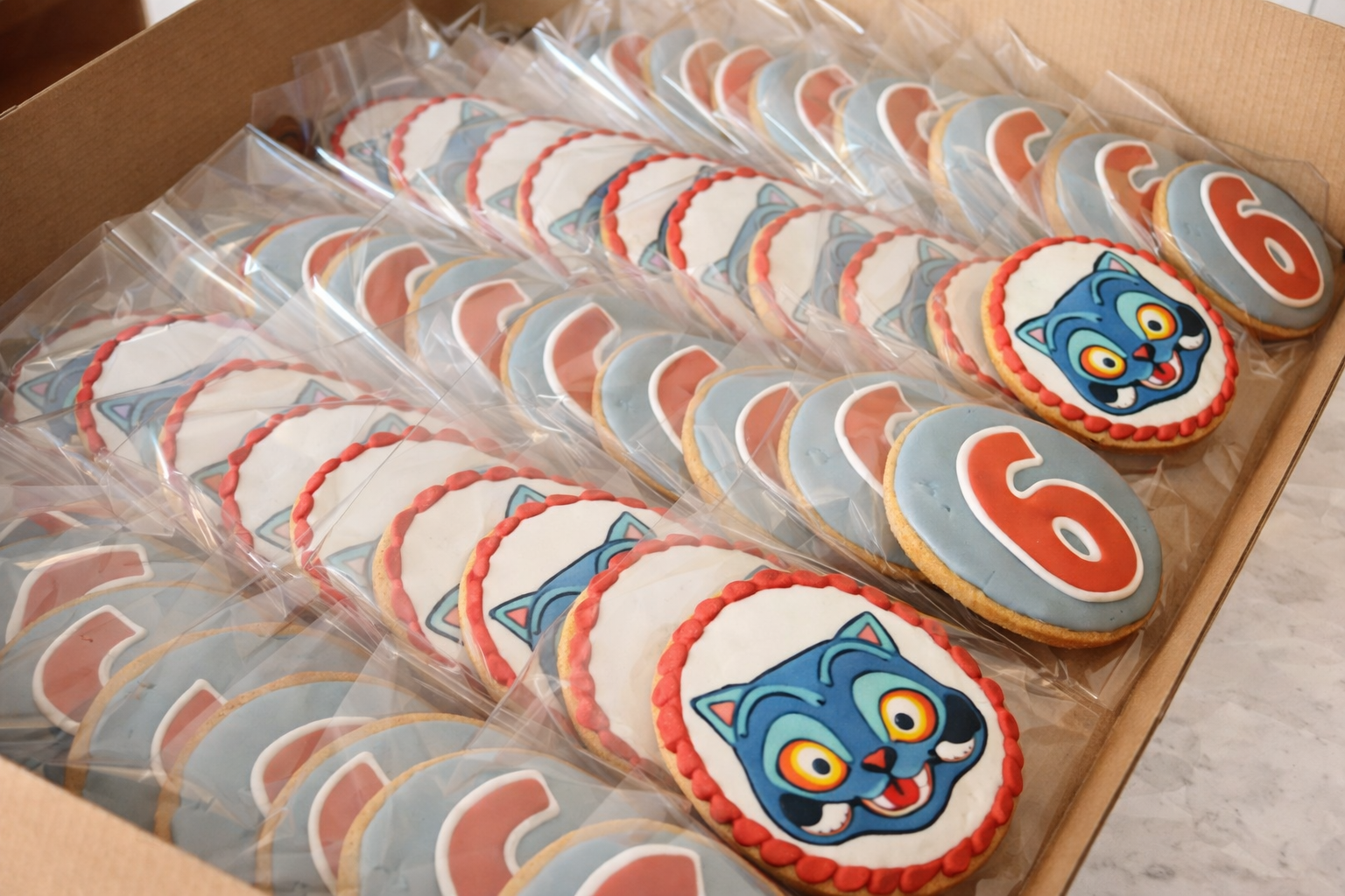 Cookies decorated with at signs, a cartoon blue cat face, and the number 6 on a light blue background, arranged in a box.