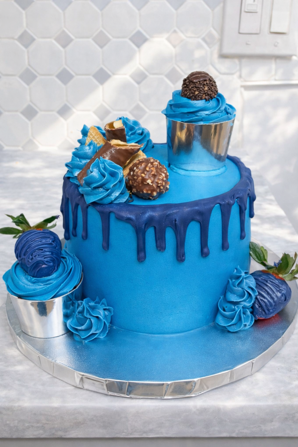A blue cake with blue drip icing, decorated with blue swirls, chocolate candies, and chocolate-covered wafer bars. The cake is on a silver cake board with some additional blue decorations and strawberries with green leaves around it.