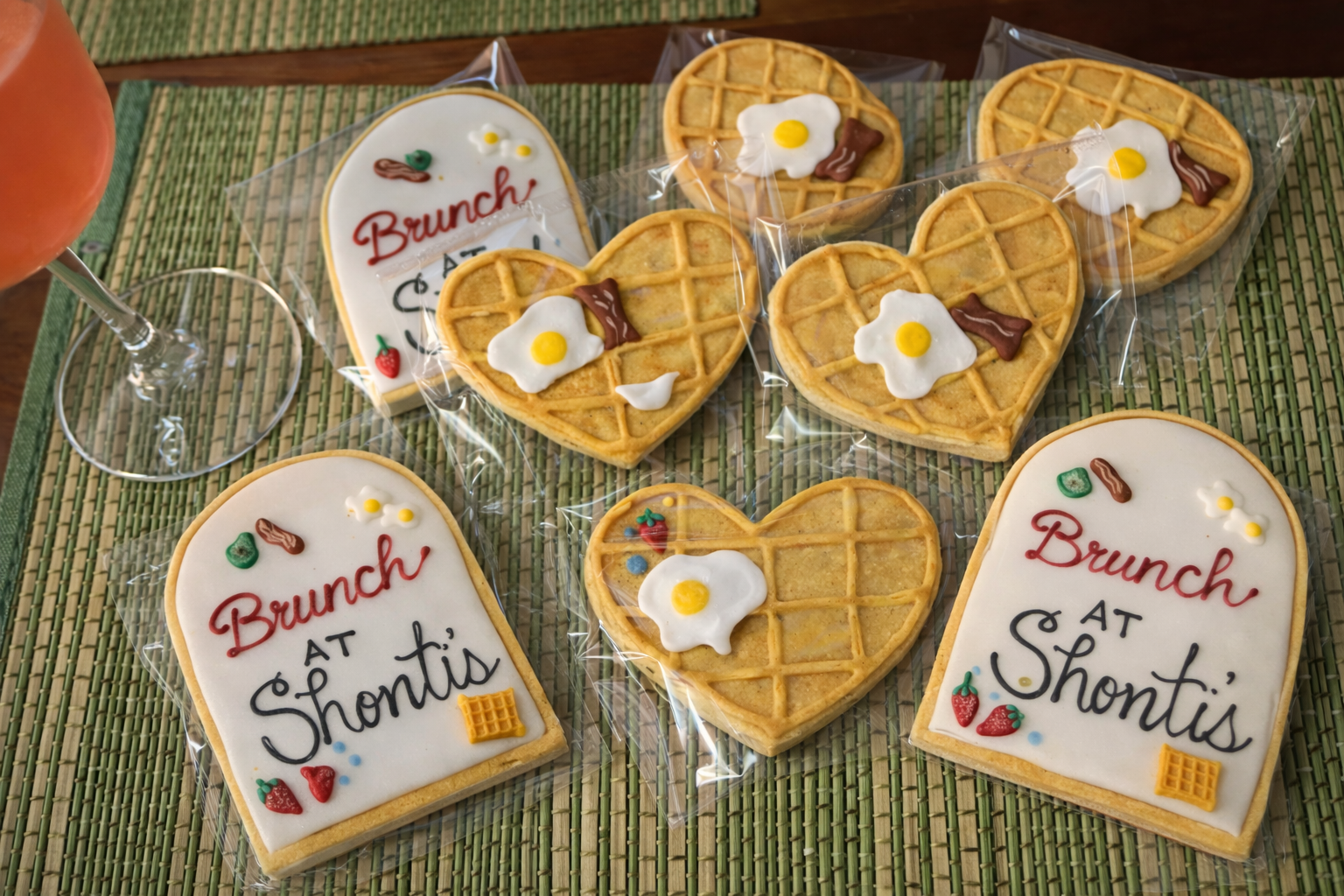 Decorated cookies with messages: 'Brunch at Shontis' and waffles with fried egg and bacon decorations, placed on a green woven placemat with a glass of pink drink.