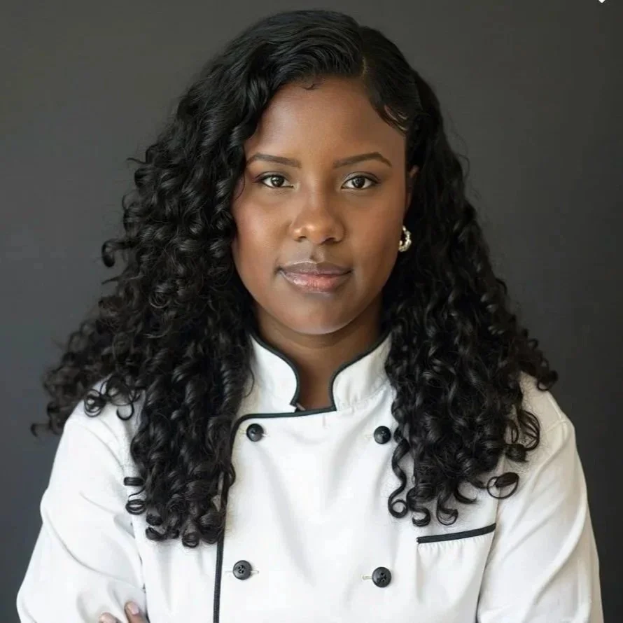 Portrait of a Black woman with long curly hair, wearing a white chef coat with black piping, standing against a dark gray background.