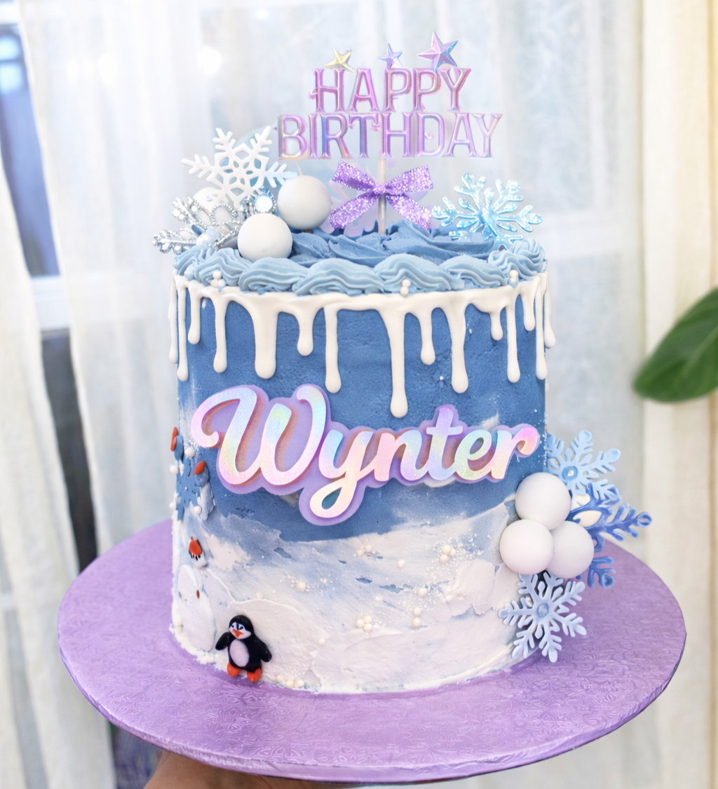 Ice blue birthday cake with white drip icing, decorated with snowflakes, white balls, and snowman figure, with a 'Happy Birthday Wynter' topper and winter theme decorations.