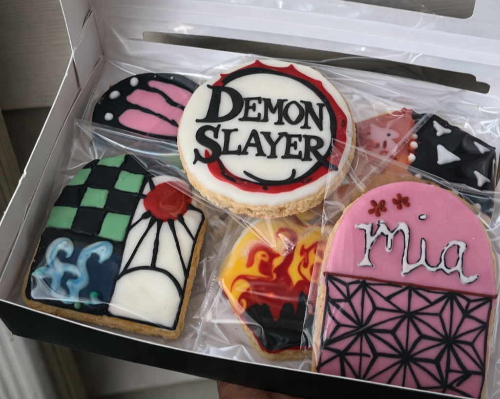 A box of decorated Halloween cookies with various designs, including one with 'Demon Slayer' written on it, a pink cookie with purple and black geometric patterns and 'Mua' written on it, and others with spider web, flames, and abstract designs.