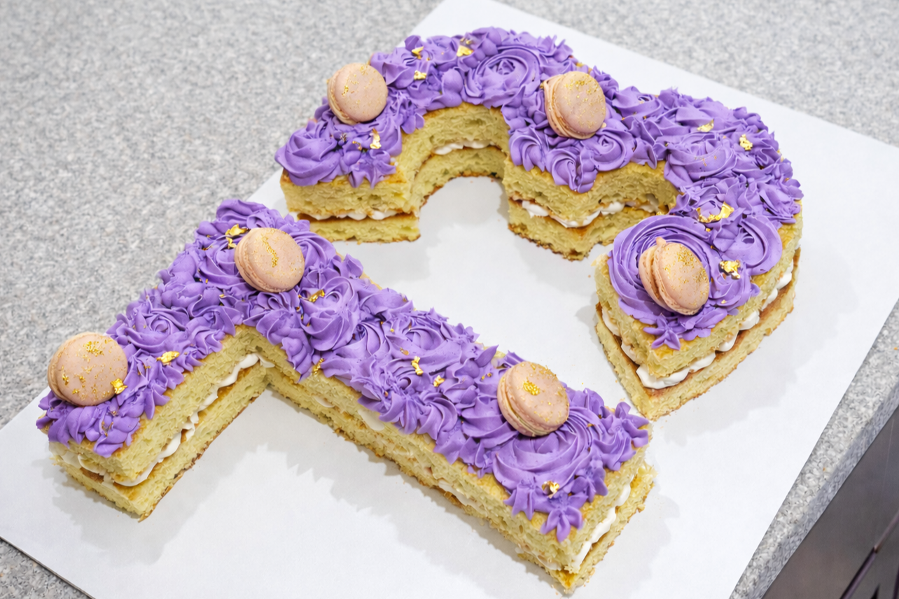 Cake decorated with purple frosting, macarons, and gold leaf, cut into a crescent shape, placed on white paper on a gray surface.