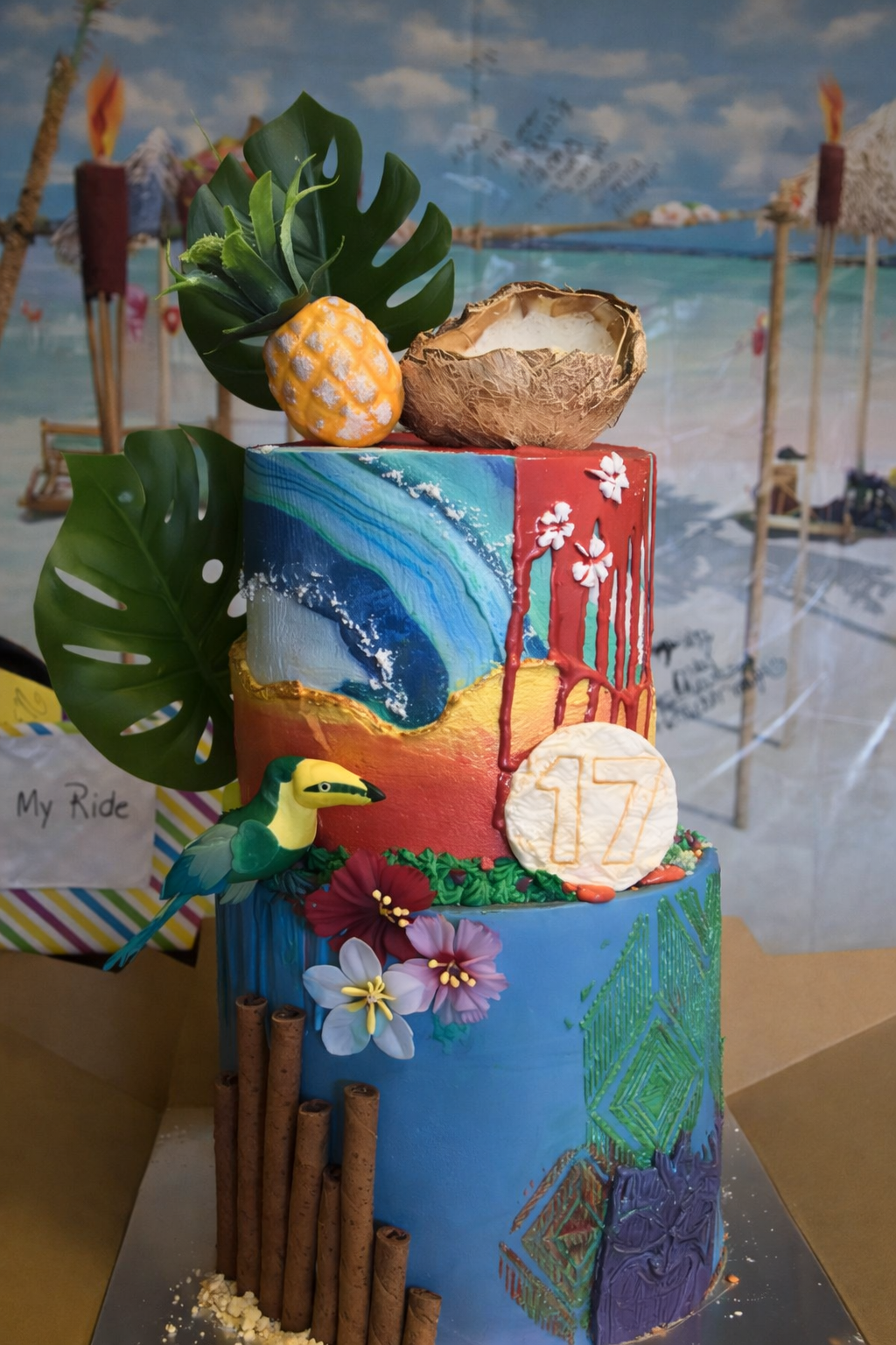 Colorful two-tiered cake with beach scene painting, tropical decorations including leaves, flowers, a pineapple, and a parrot figurine, set against a beach-themed backdrop.