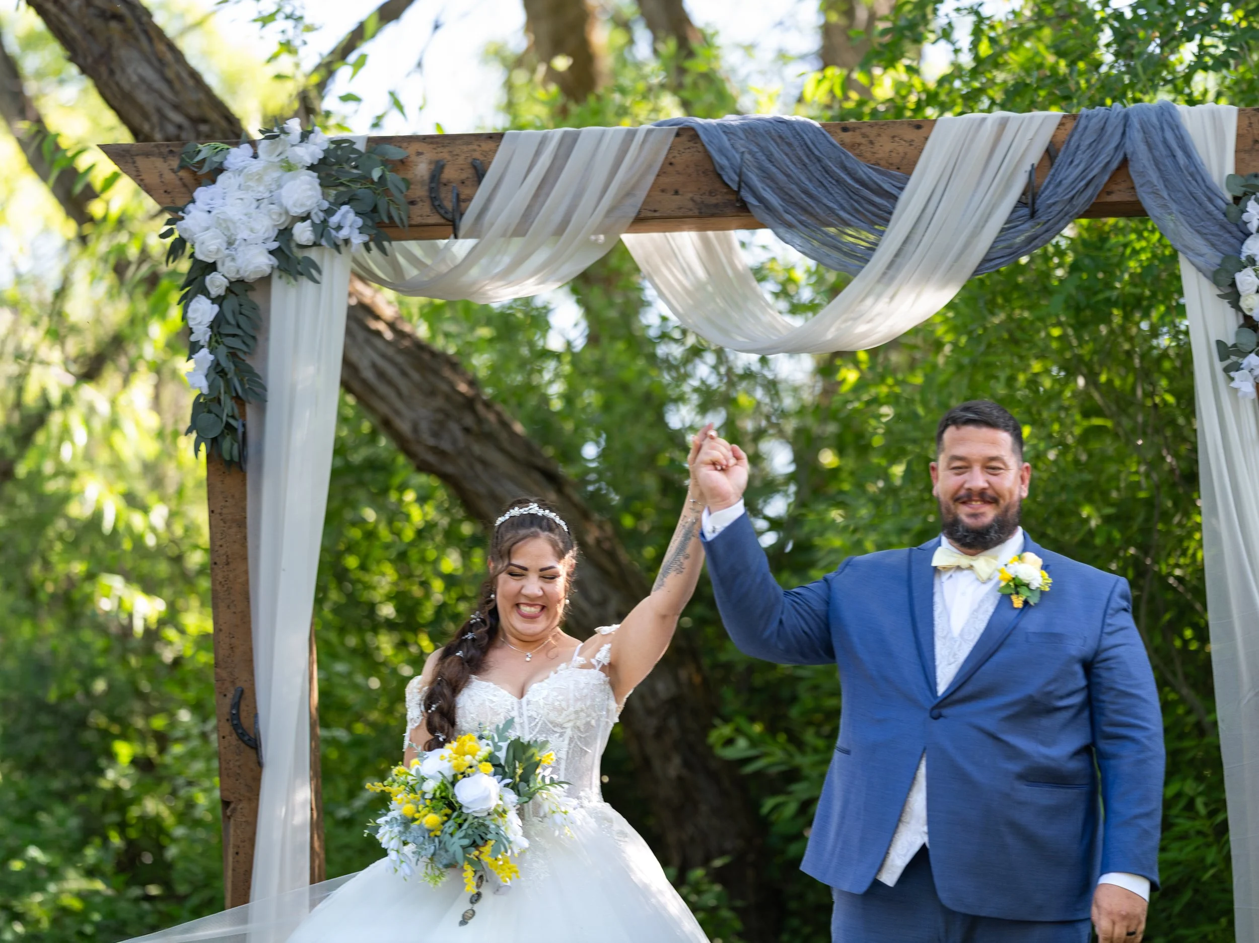 A bride and groom celebrating outdoors at their wedding, holding hands and smiling, with a decorated wooden arch featuring white and gray fabric and white flowers.