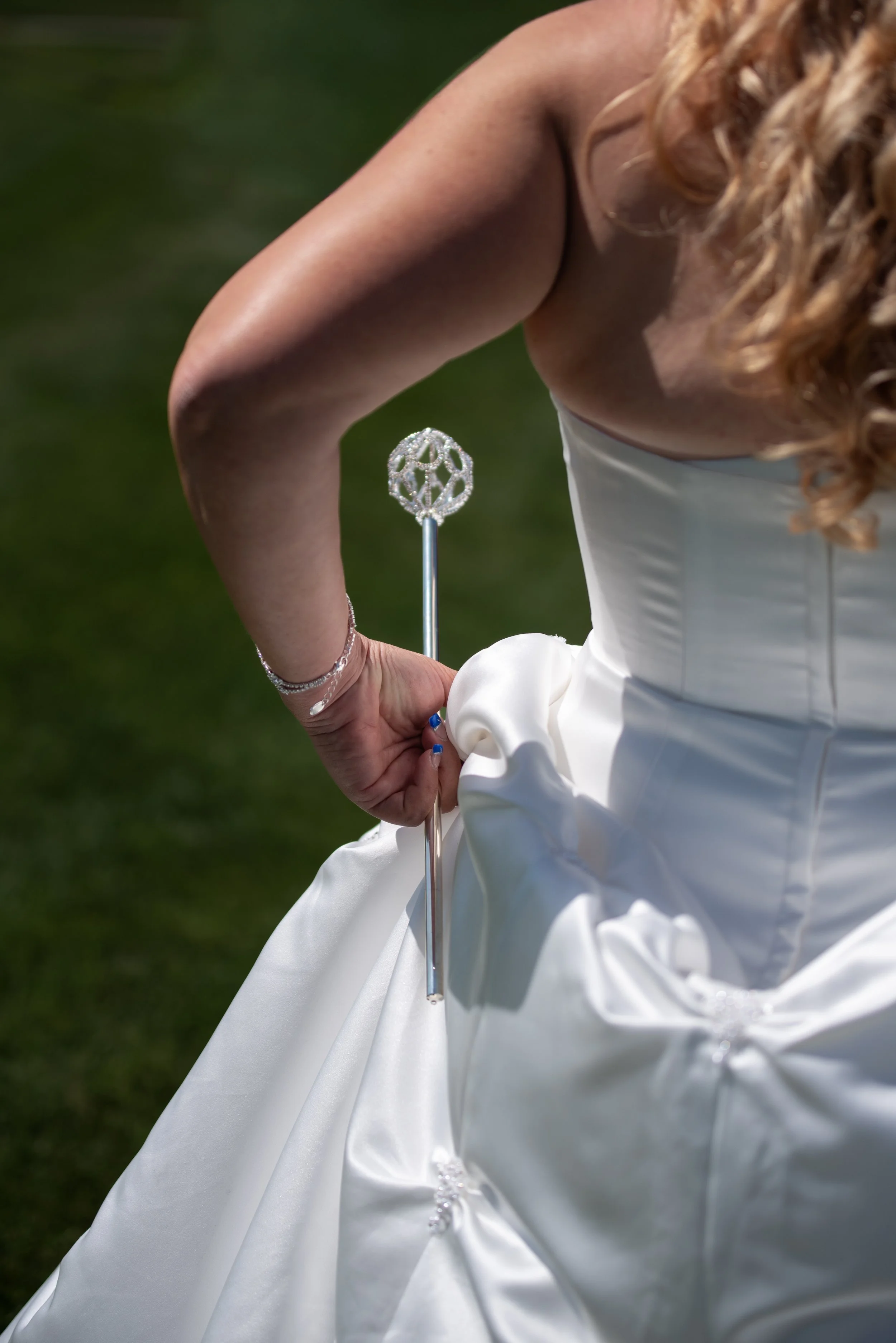 Close-up of a woman in a white wedding dress holding a decorative sparkly wedding stick, with focus on upper body and part of the dress, outdoors on green grass.