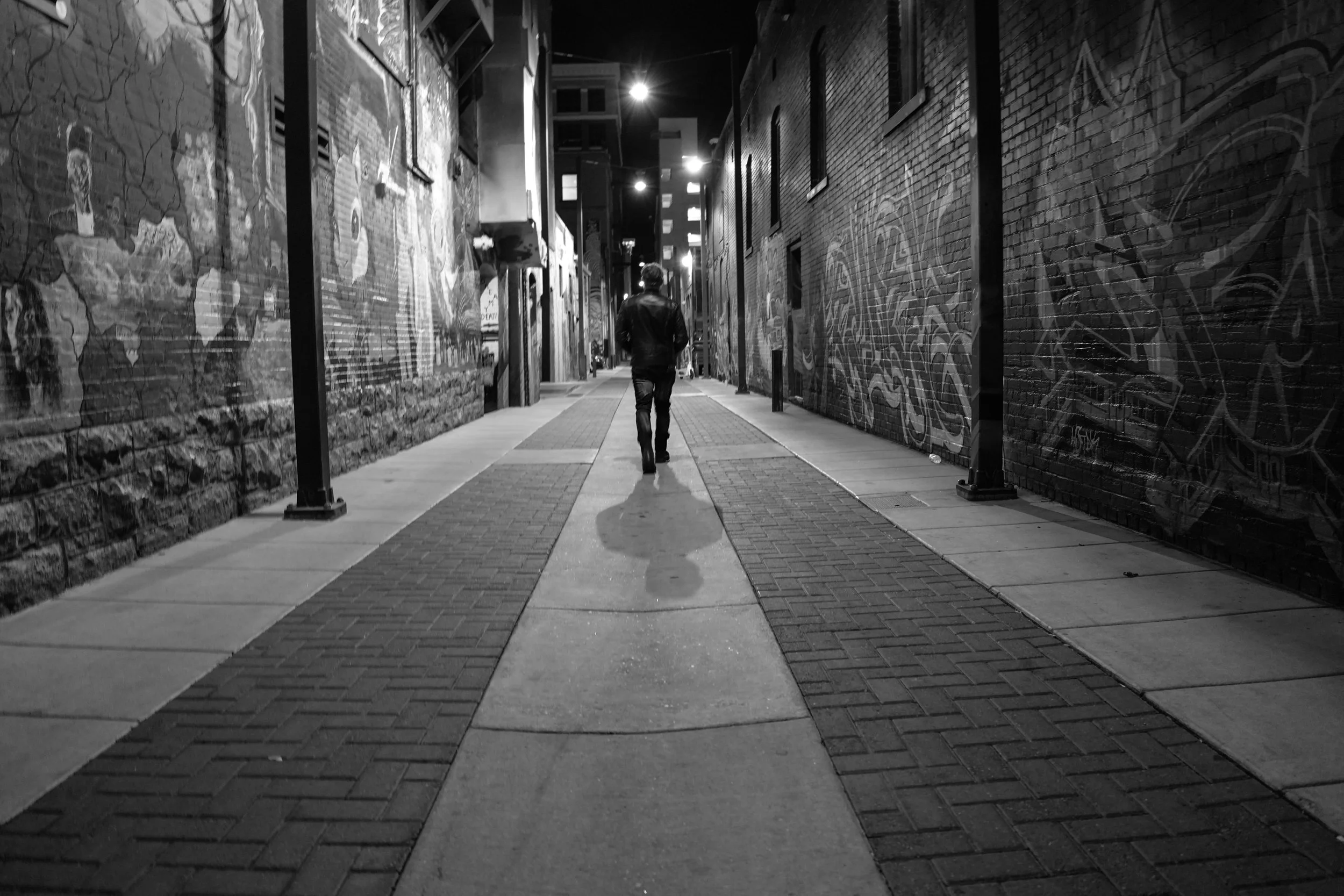 A person walks down a city alley at night, with graffiti-covered brick walls on both sides, illuminated by streetlights.