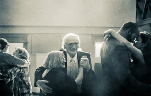 Older couple dancing closely among other couples at a social gathering in a hall.