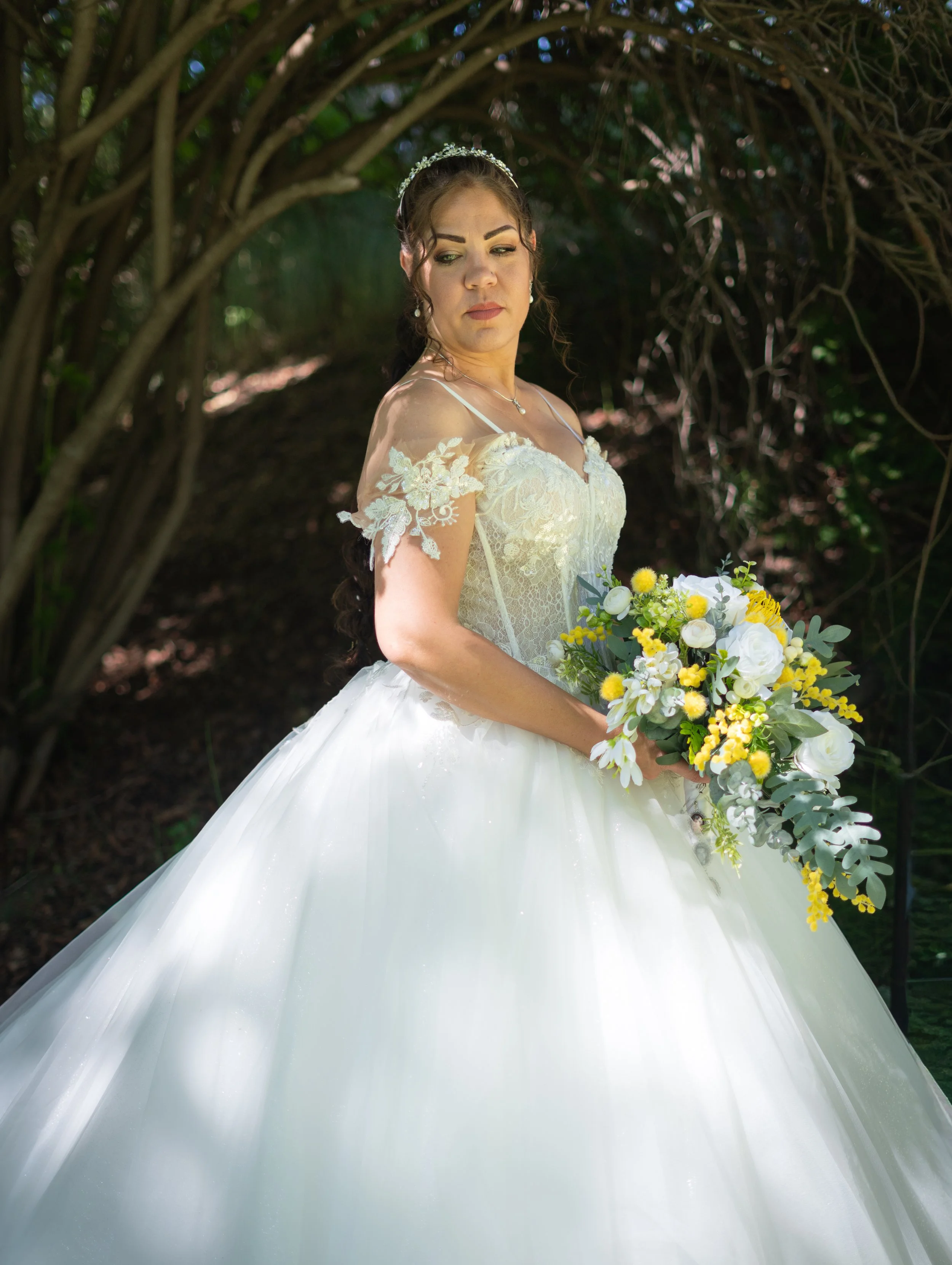 A bride in a white wedding dress with lace and embroidery, holding a bouquet of yellow and white flowers, standing outdoors under green trees.