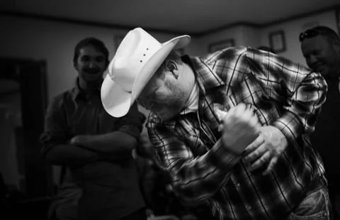A man wearing a cowboy hat and plaid shirt flexing his arm while two other men watch and smile in the background.