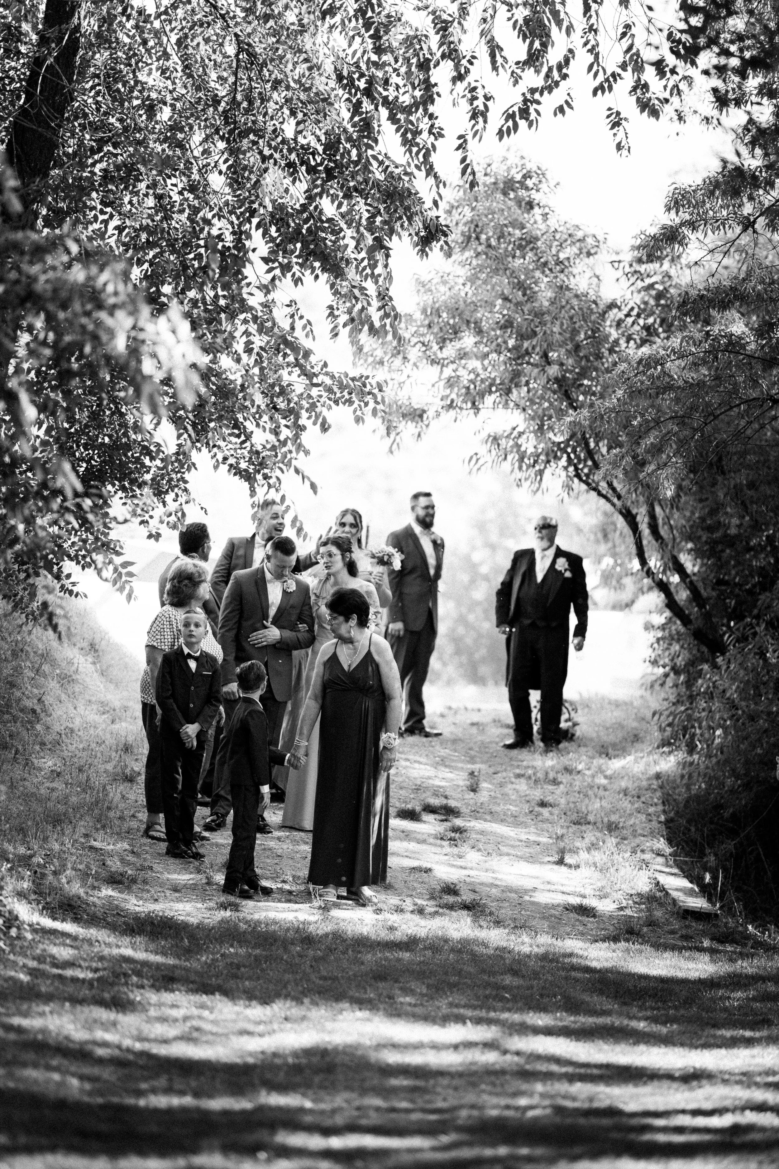 A black and white photograph of a wedding procession walking along a wooded path, with several people dressed in formal attire, including a bride and groom, surrounded by trees and natural scenery.