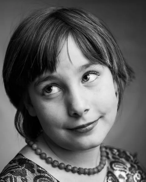 Black and white portrait of a young girl with short hair, wearing a patterned top and a beaded necklace, looking to the side with a slight smile.