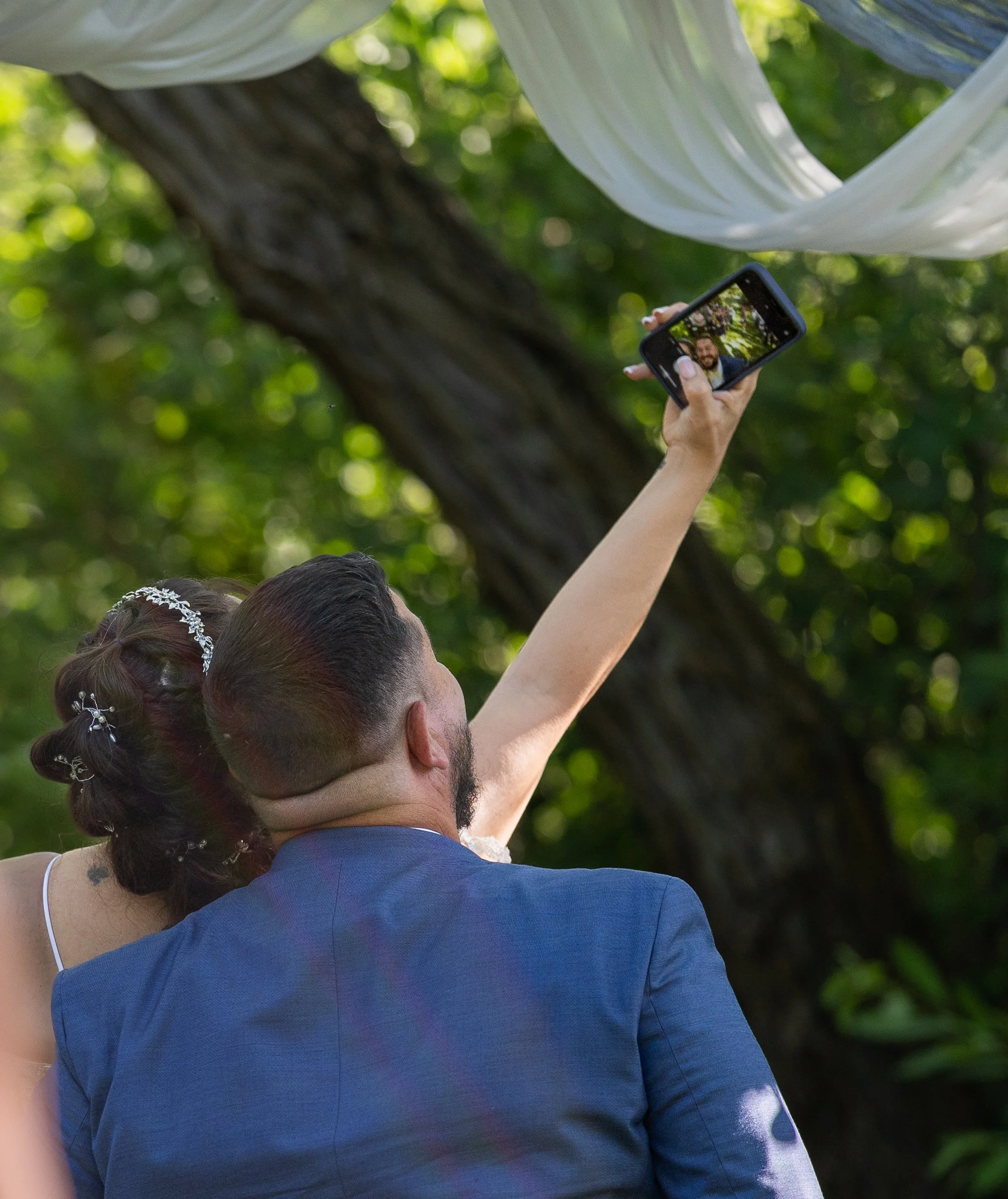 A man and woman taking a selfie outdoors during a wedding celebration with trees and greenery in the background.
