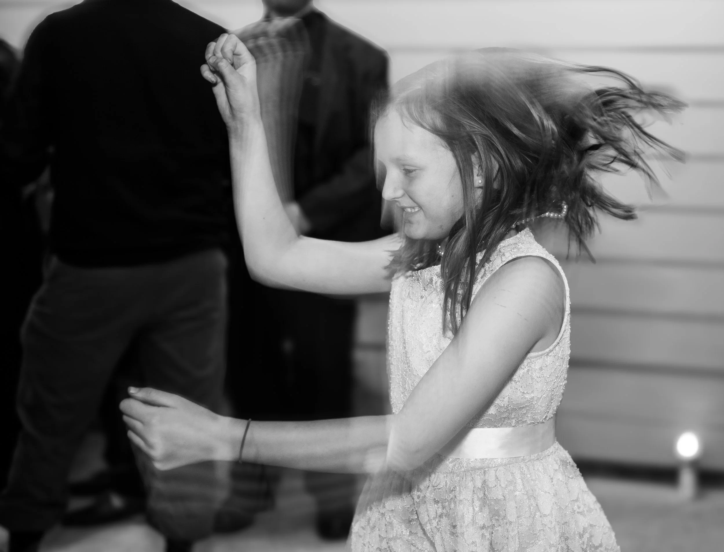 A young girl with long hair dancing with her eyes closed, smiling, wearing a laced dress, with hair flipping to the right, in black and white.