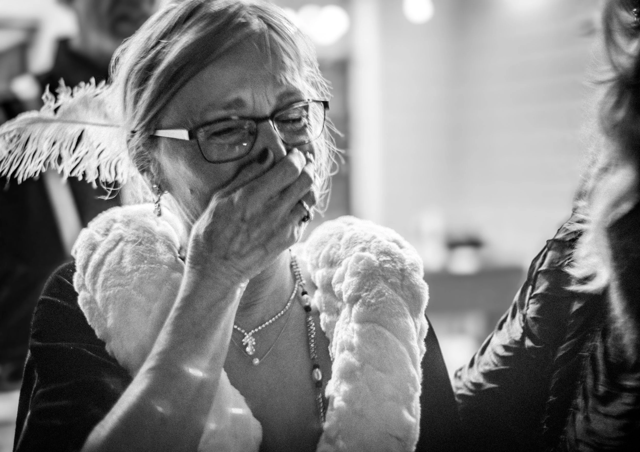 An elderly woman with glasses and earrings, wearing a necklace and a fur-lined coat, is covering her mouth with her hand and appears to be emotional or surprised. She is in a social setting with other people around.