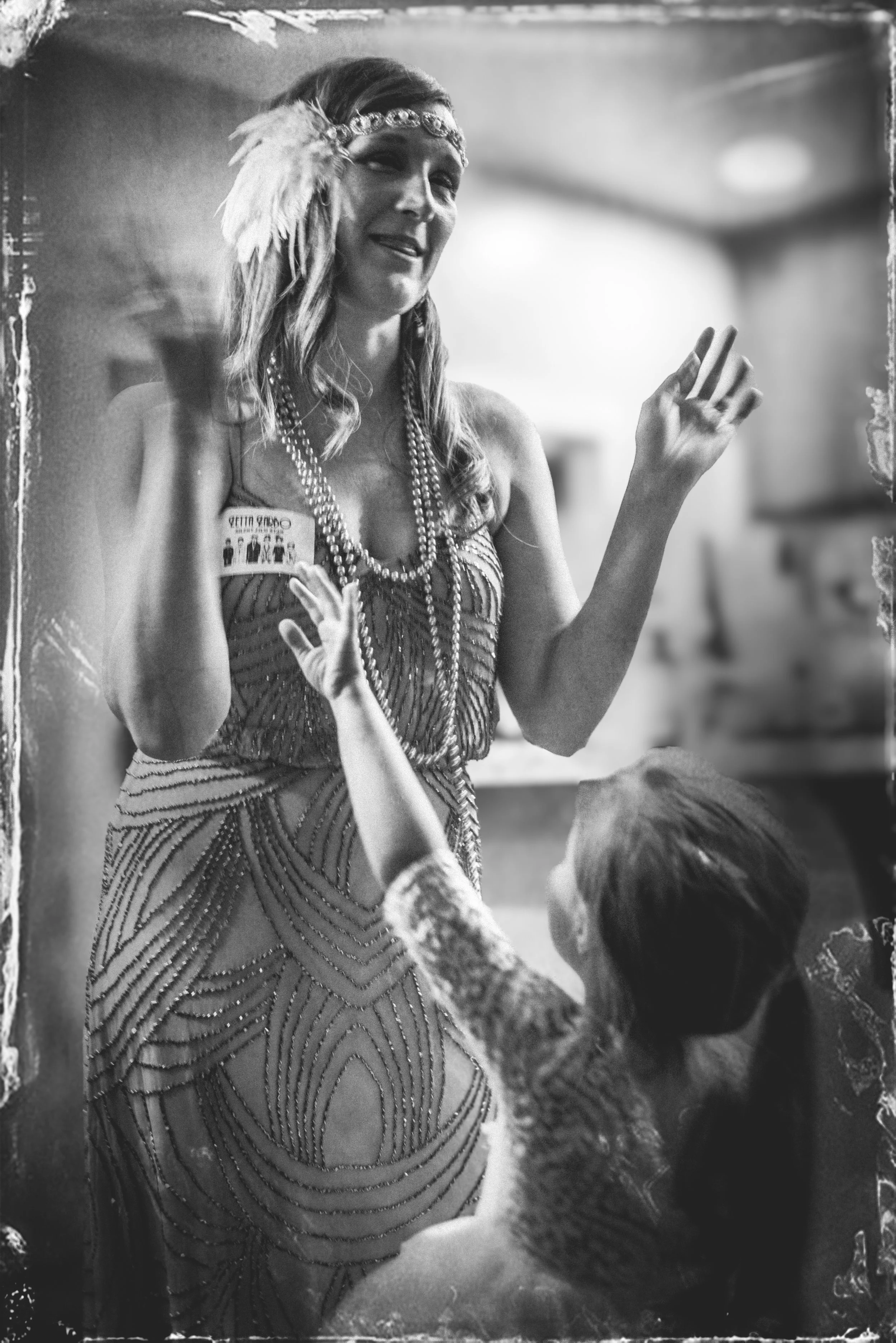 A woman wearing 1920s-style flapper dress, headband with feathers, and long pearls, is smiling and waving at a young girl with short hair, who is reaching out to her.