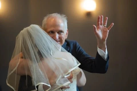 A man and a woman in wedding attire hugging, with the man waving and smiling at the camera.