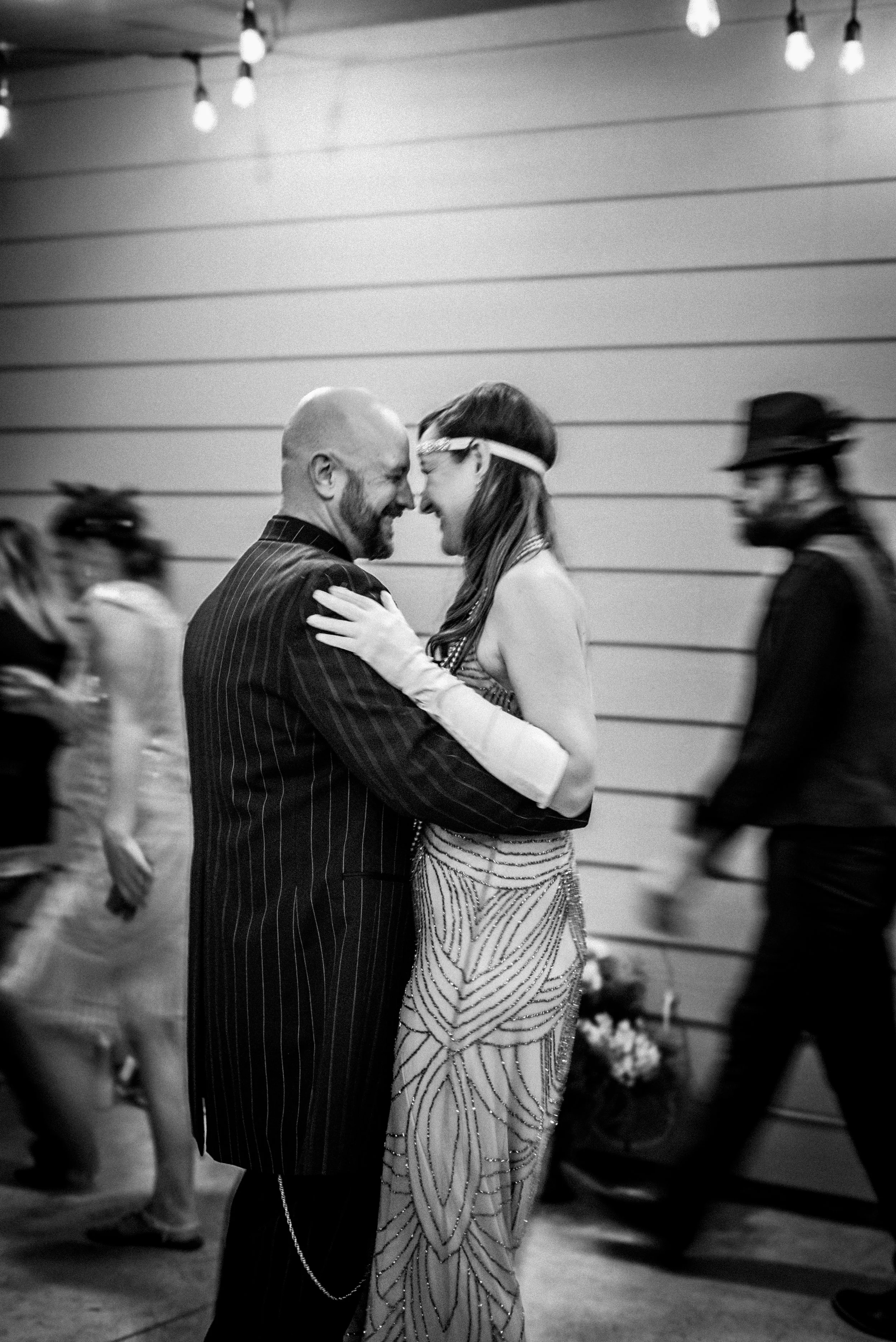 A man and woman dancing closely and smiling at each other at a party or dance event, with other guests in the background.
